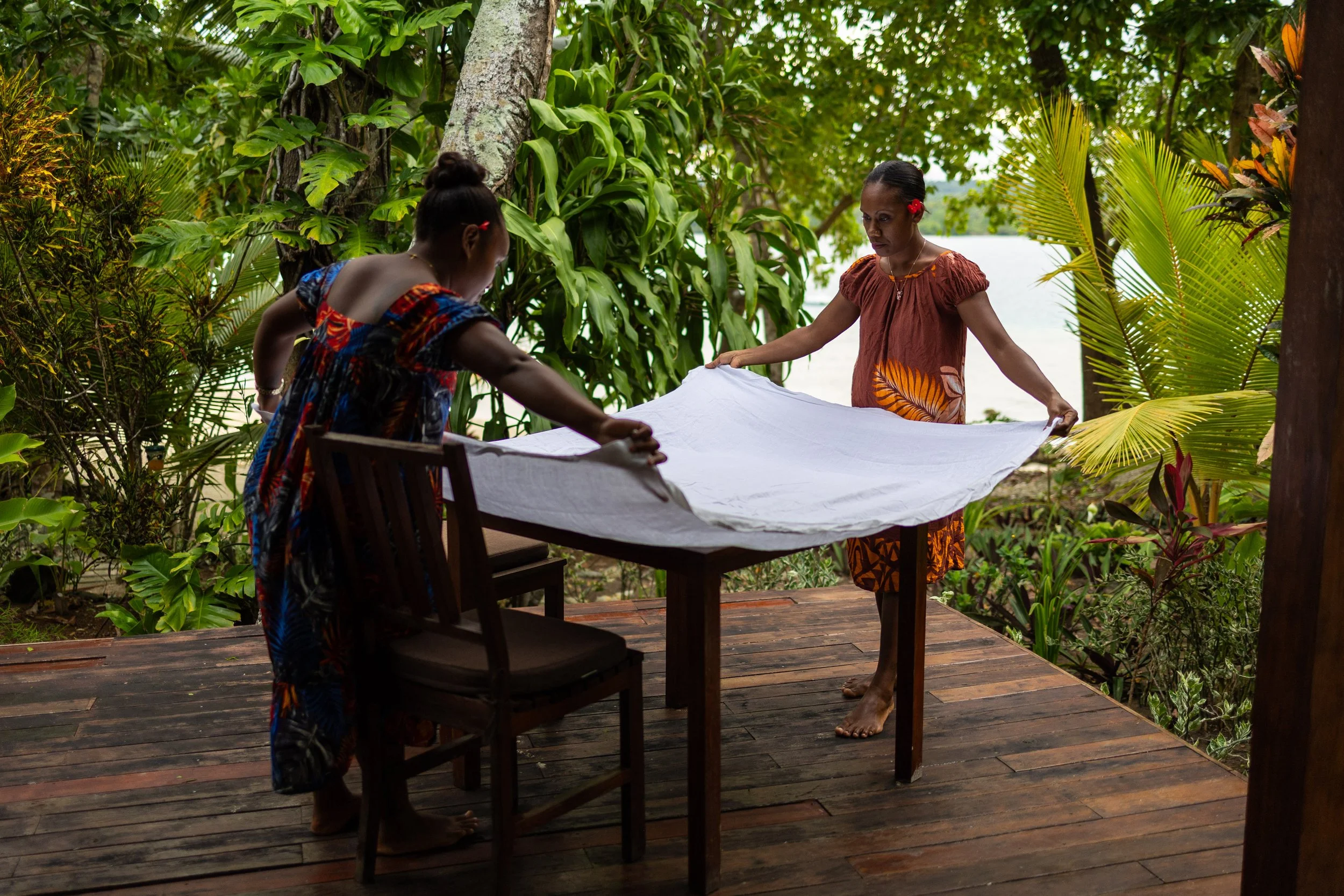 Two women in traditional clothing setting up a white cloth on a wooden table outdoors surrounded by lush green tropical plants.