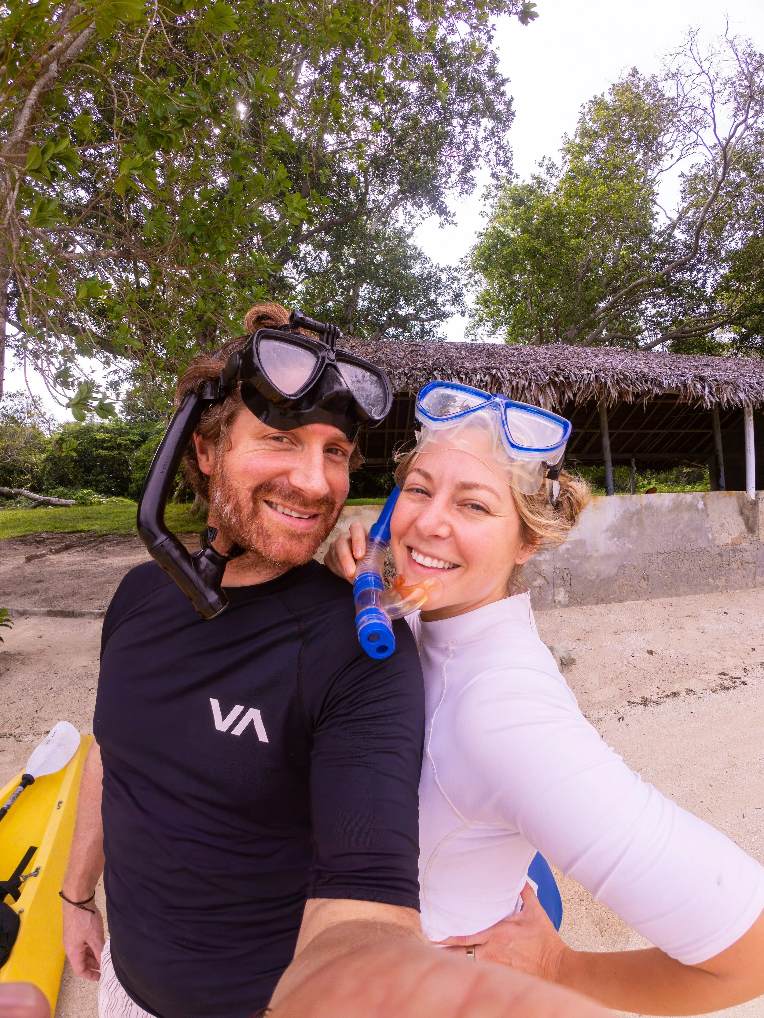 A smiling man and woman in snorkeling gear taking a selfie on the beach. The man is wearing a black rash guard and a snorkel mask; the woman is wearing a white rash guard and a snorkel mask. Behind them are trees, a thatched hut, and sandy beach.