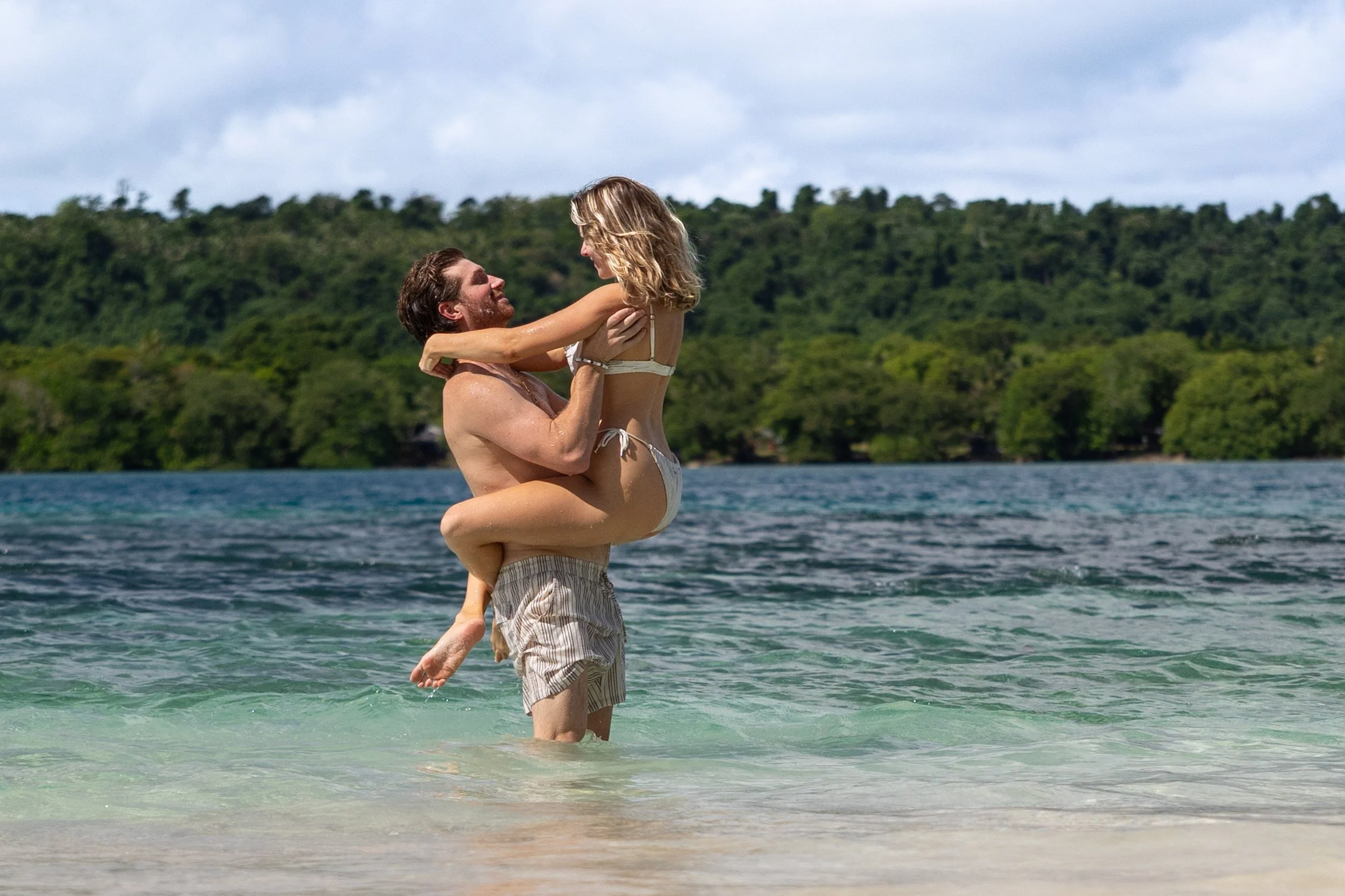 A man lifting a woman in the water at the beach, with a scenic landscape of green hills and cloudy sky in the background.
