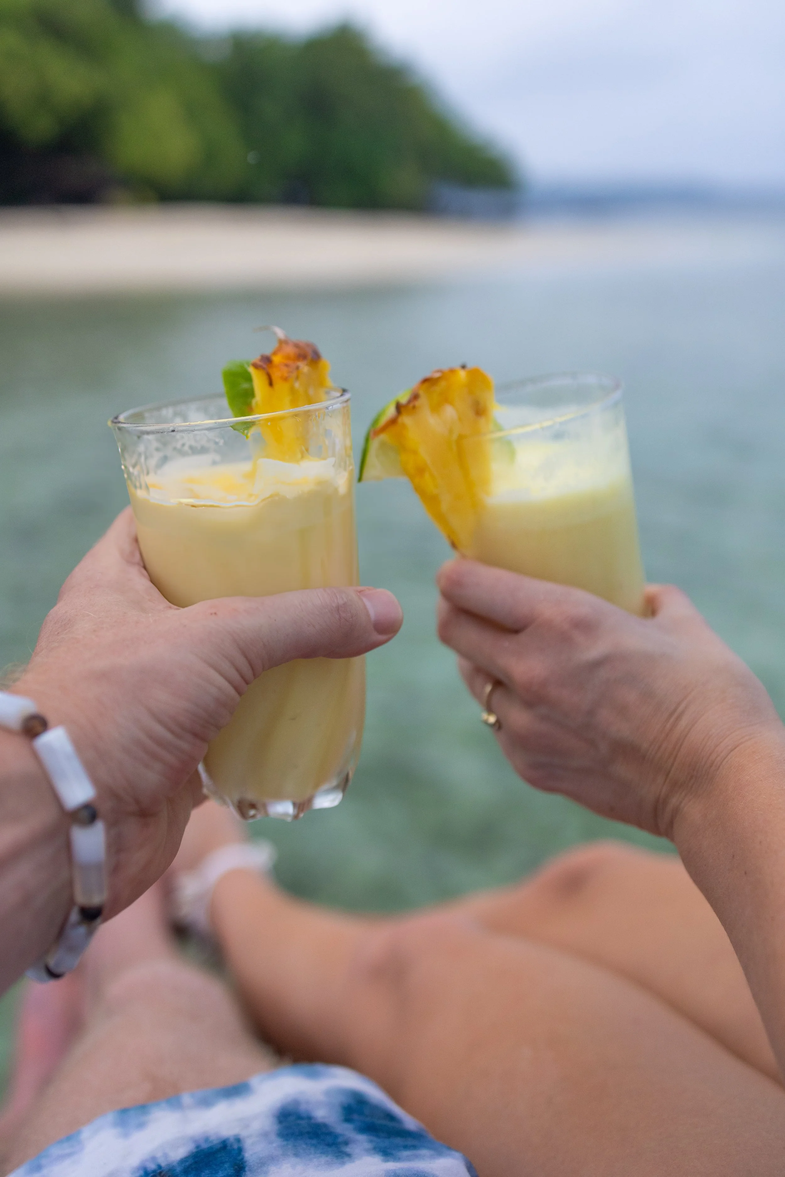 Two people holding tropical drinks with pineapple slices against a beach and ocean background.