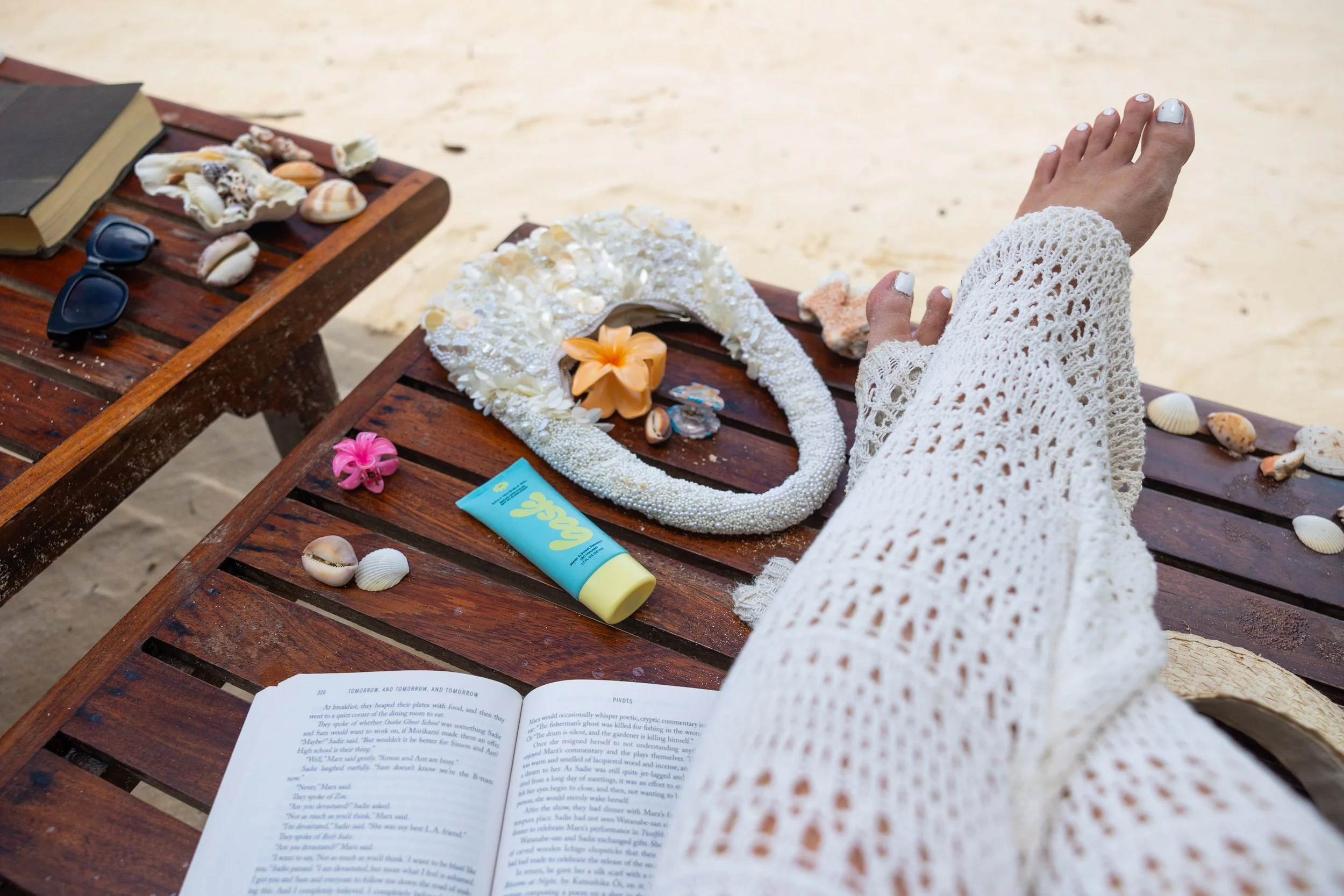 Person in a white crochet cover-up lying on a wooden beach chair, with seashells, flowers, and beach accessories scattered on a wooden table nearby at the beach.