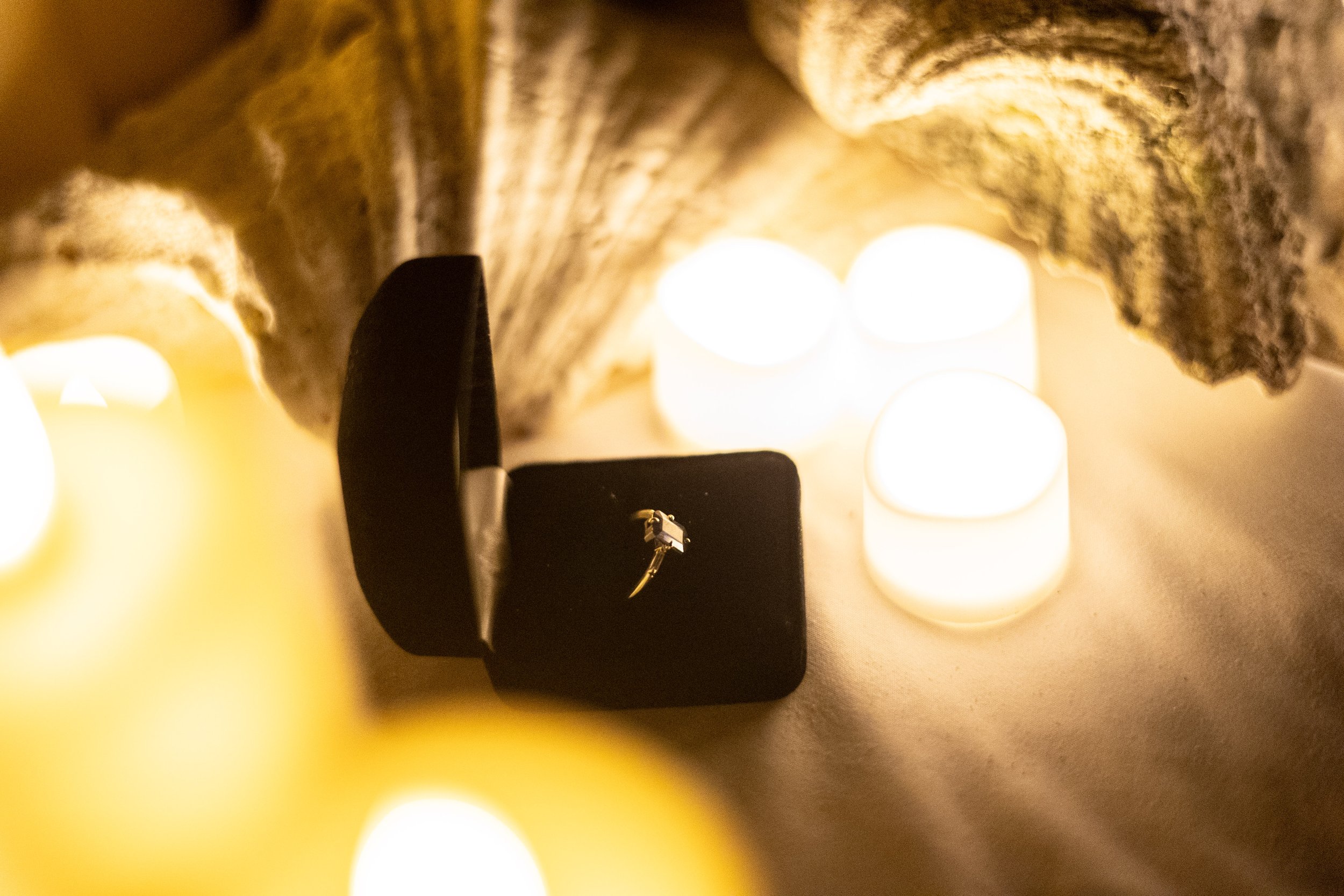A small diamond ring is displayed inside an open black ring box, surrounded by tealight candles creating a warm, romantic atmosphere.