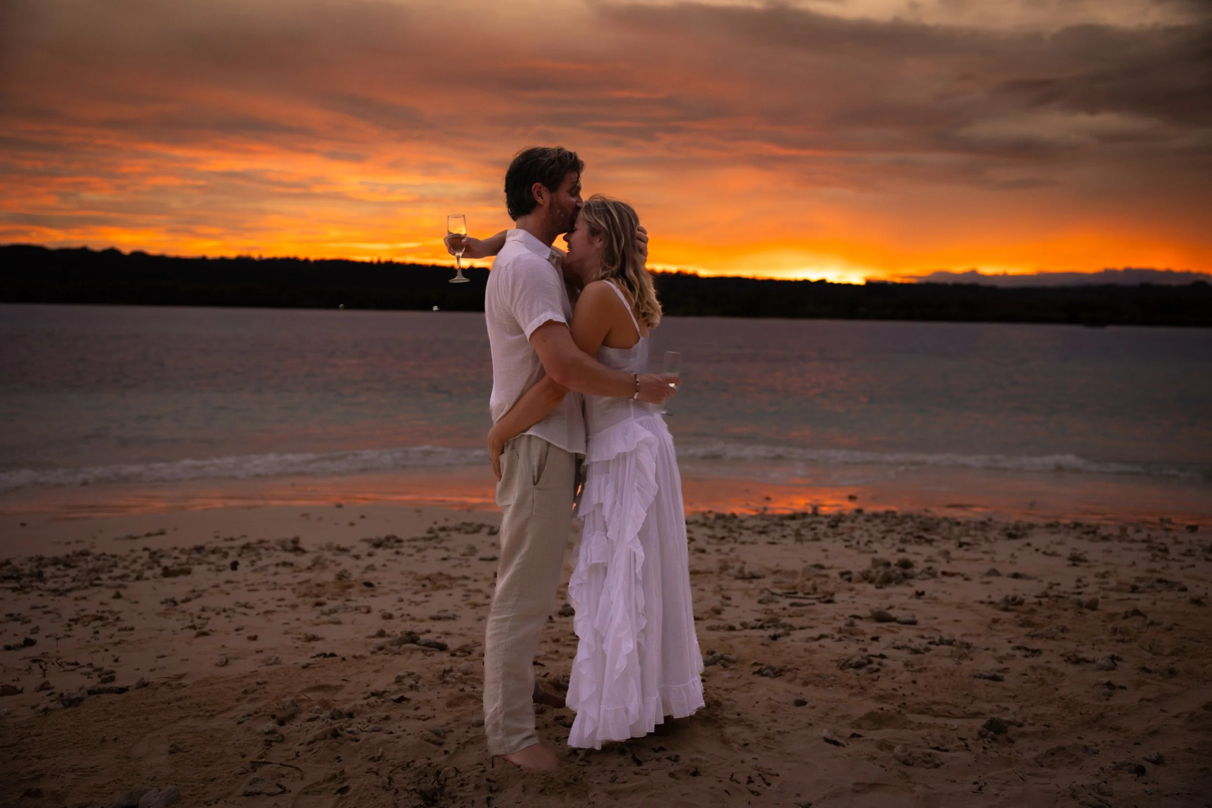A couple embracing on a beach at sunset, holding wine glasses, with a colorful sky and calm water in the background.