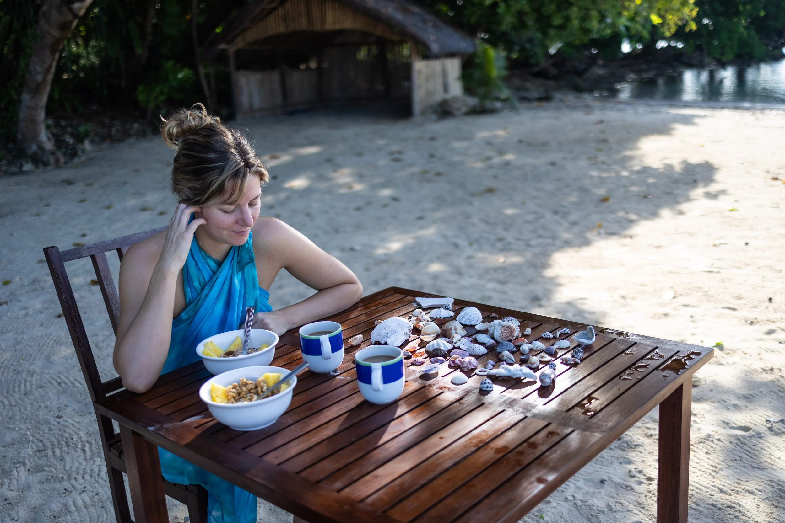 A woman in a blue dress sitting at a wooden table on a beach, with bowls of cereal and cups of coffee, looking at seashells on the table near the water.