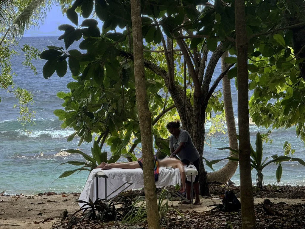 A person receiving a massage on a massage table under a tree on a beach with ocean waves in the background.