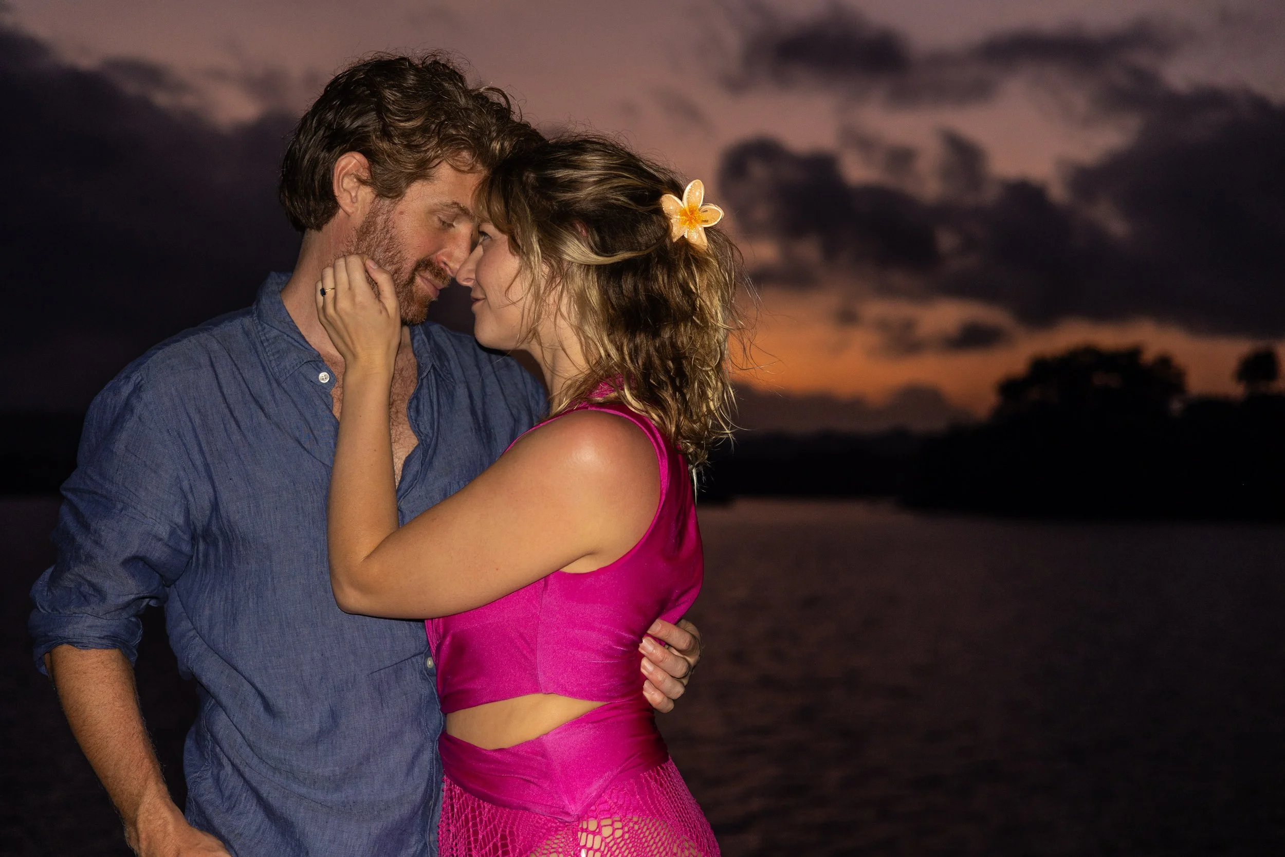 A romantic couple standing close together outdoors at sunset with dark clouds in the sky, the woman wearing a pink dress with a flower in her hair, and the man wearing a blue shirt. They are touching foreheads and smiling gently at each other.