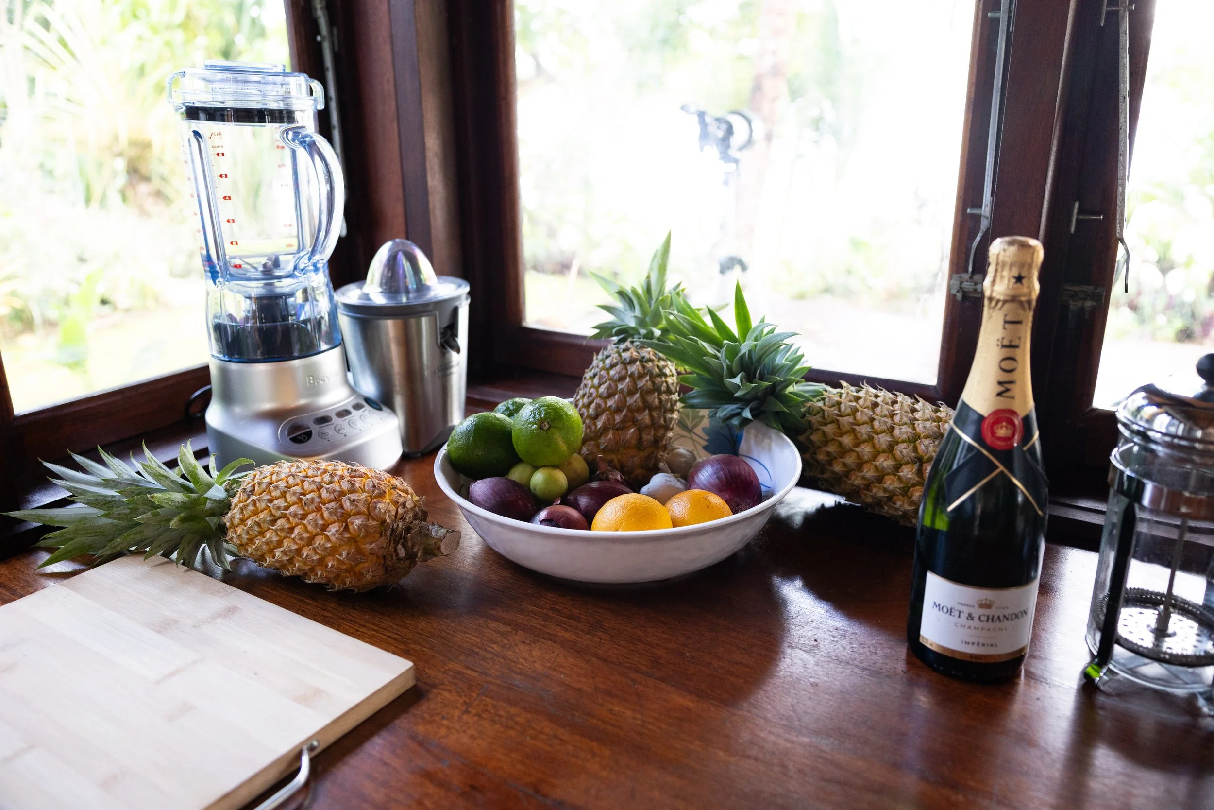 A wooden kitchen counter with pineapples, a bowl of mixed fruits, a blender, a citrus juicer, a bottle of Moët & Chandon champagne, and a glass jar near a window with outdoor garden view.