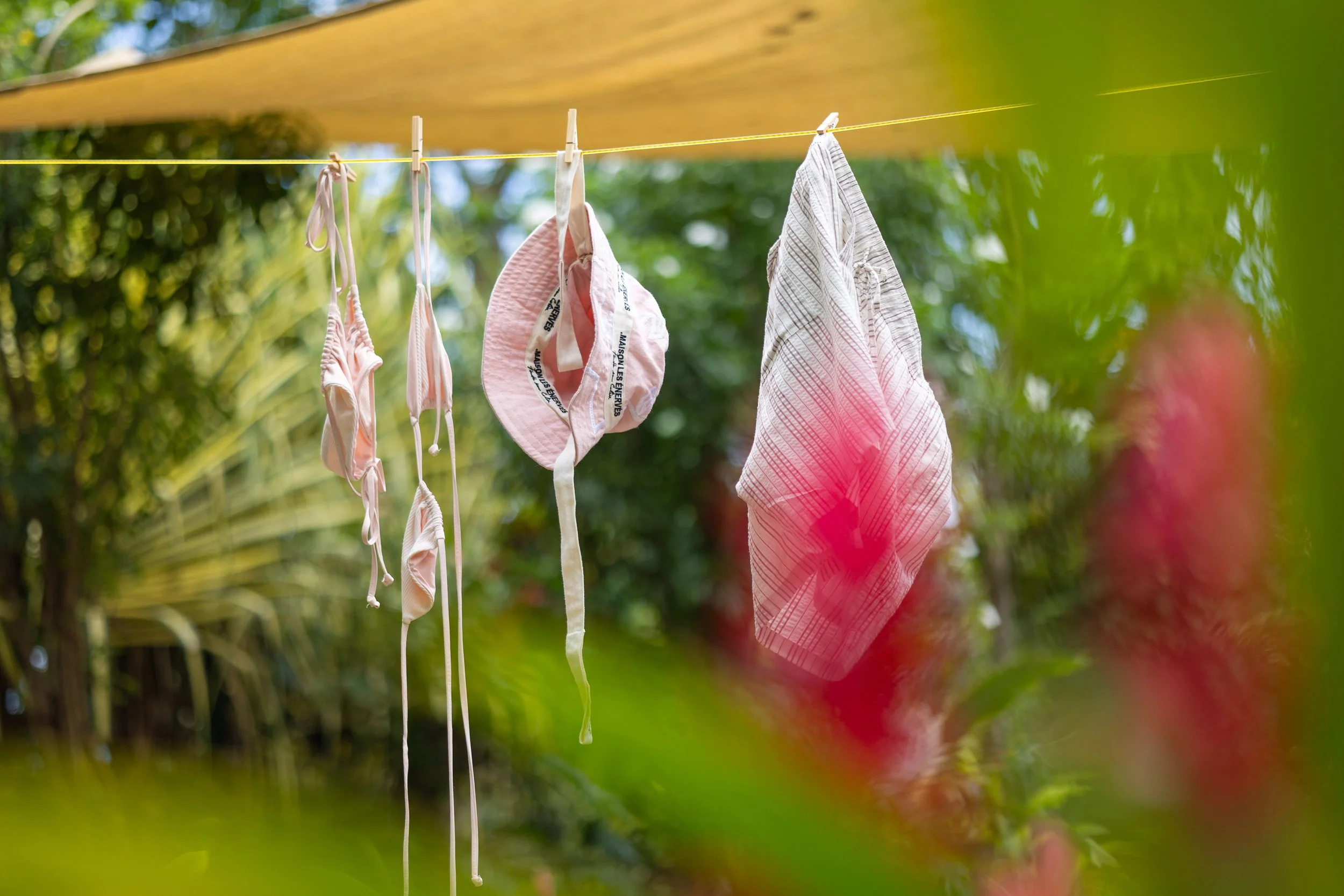 Clothes, including a bikini, a hat, and a towel, hanging on a clothesline outdoors with green foliage in the background.