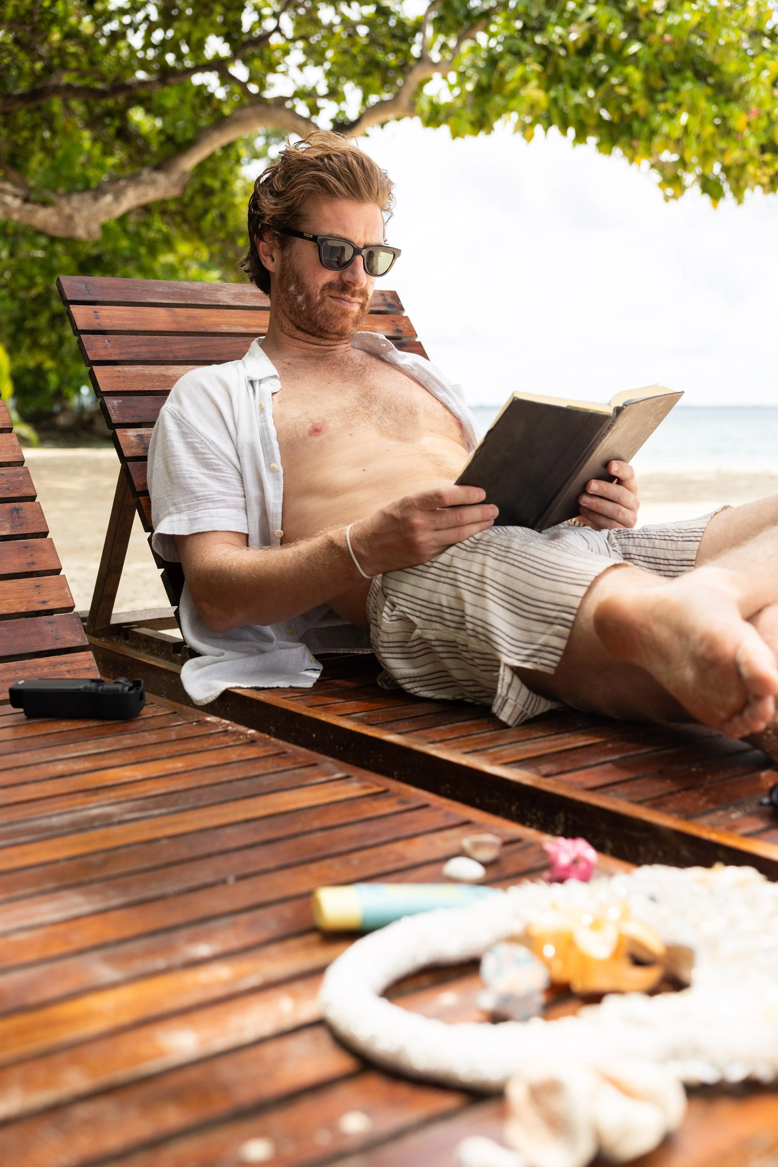 A man with sunglasses, open shirt, and shorts relaxing on a wooden lounge chair under a tree on the beach, reading a book.