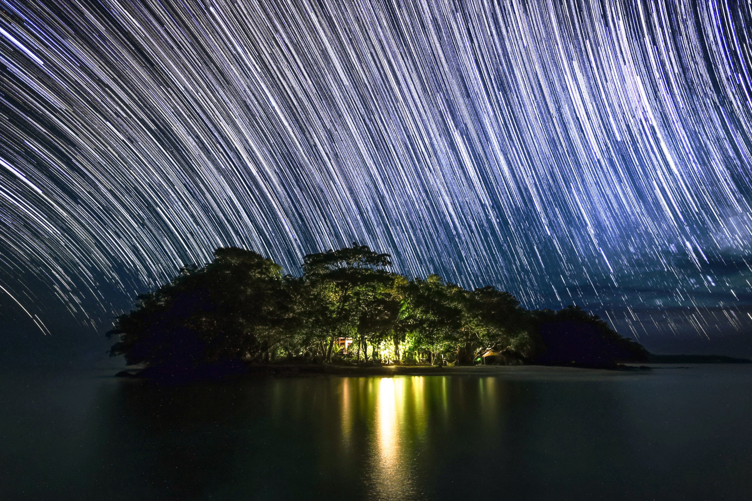 Night sky filled with star trails over a small island with trees, illuminated by a light source, reflected in calm water.