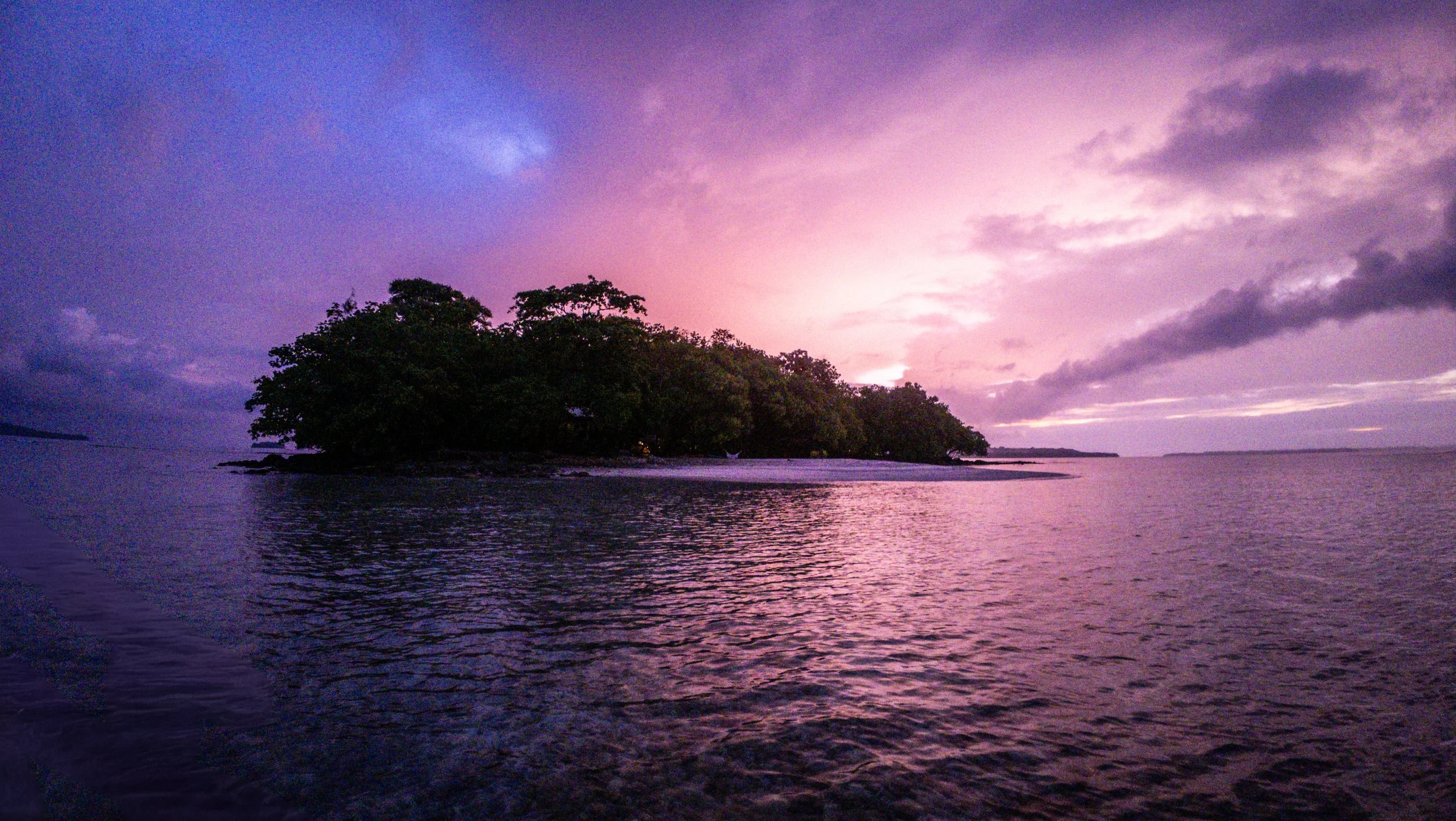 A tropical island with lush green trees surrounded by water during sunset with a purple and pink sky.