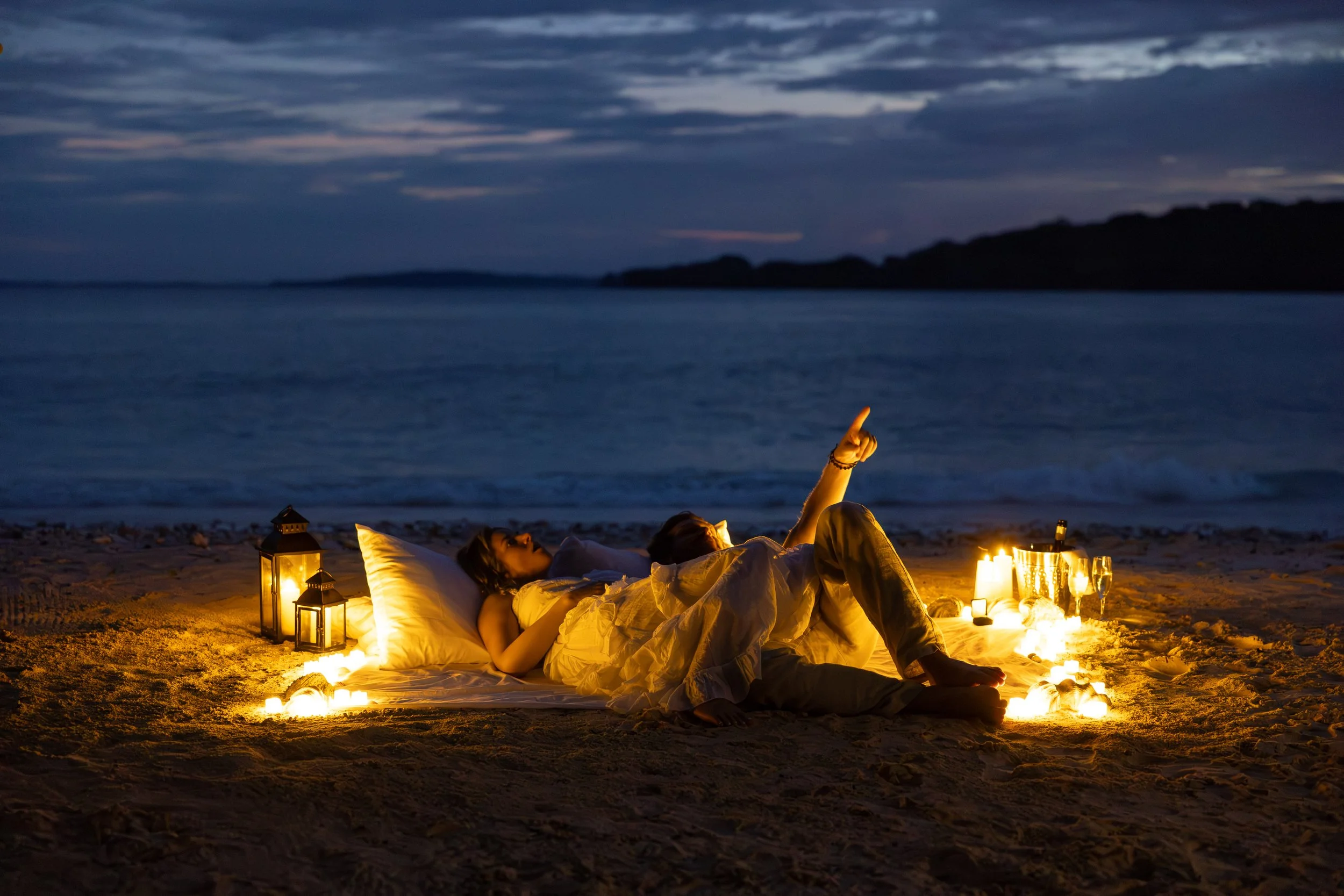 A couple relaxing on a beach at night, lying on a blanket surrounded by candles, lanterns, and glasses, with the ocean and a cloudy sky in the background.
