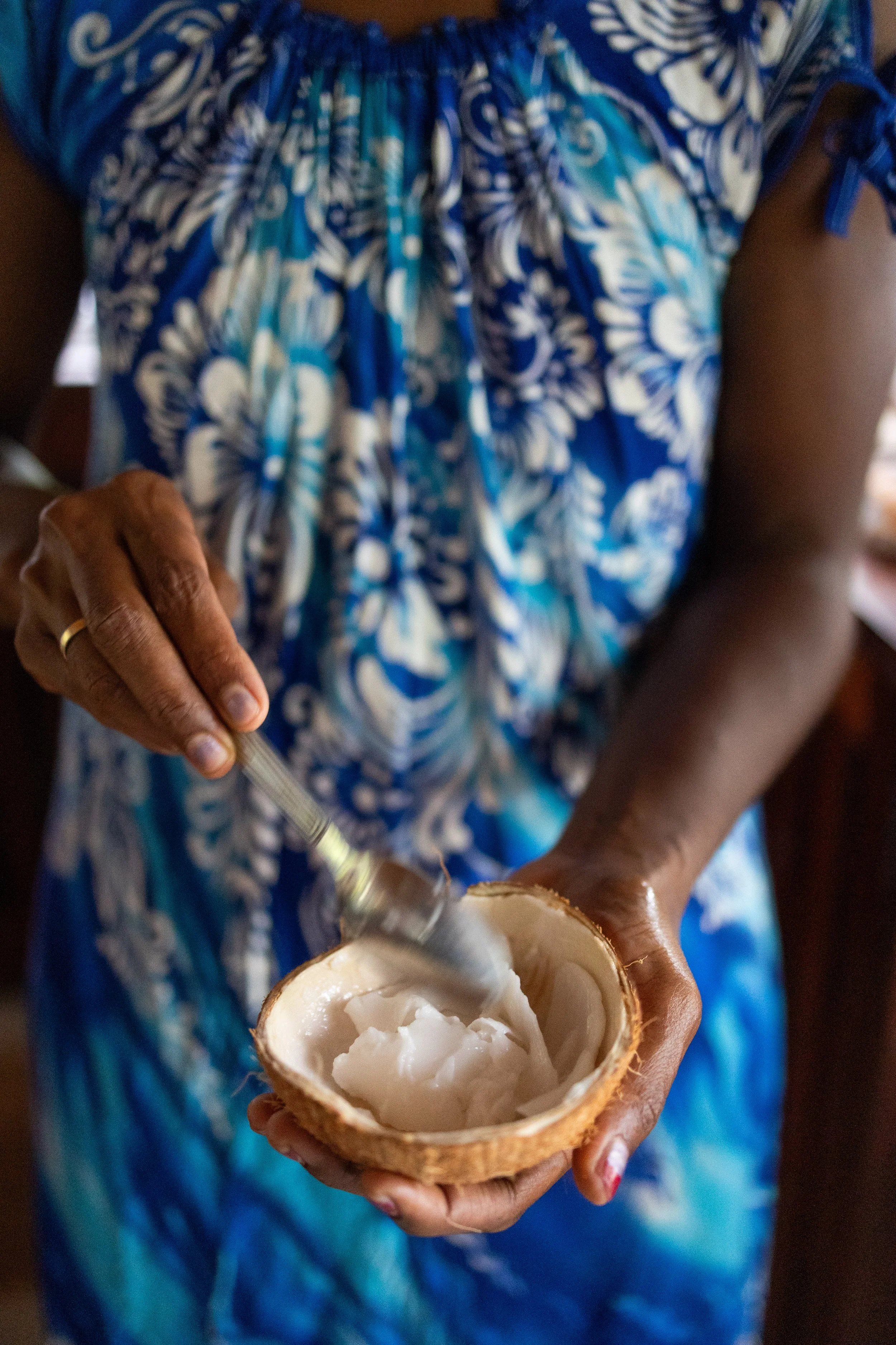 Person in blue patterned dress scooping coconut from a halved coconut shell.