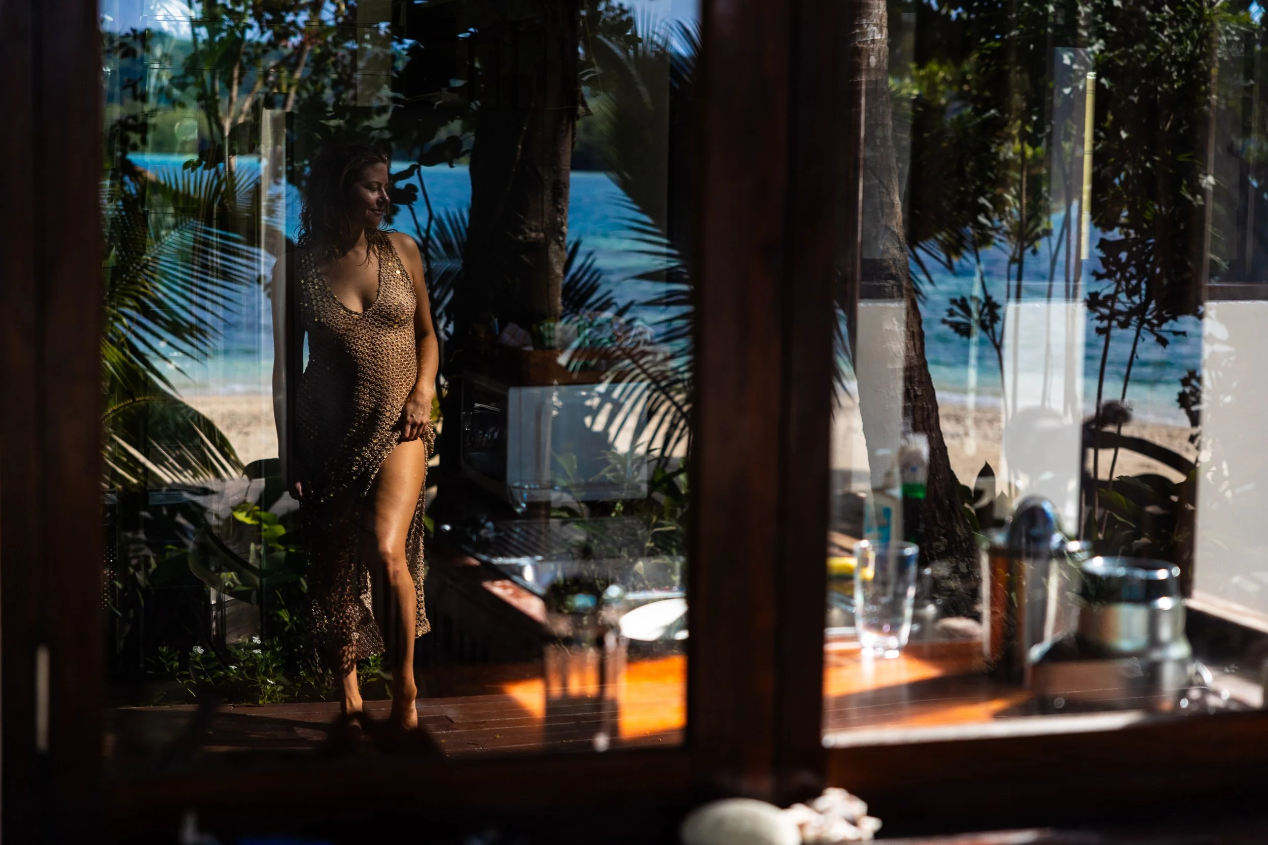 Woman in a brown, see-through dress standing inside a room, viewed through a glass window with reflections of tropical trees and a beach scene outside.