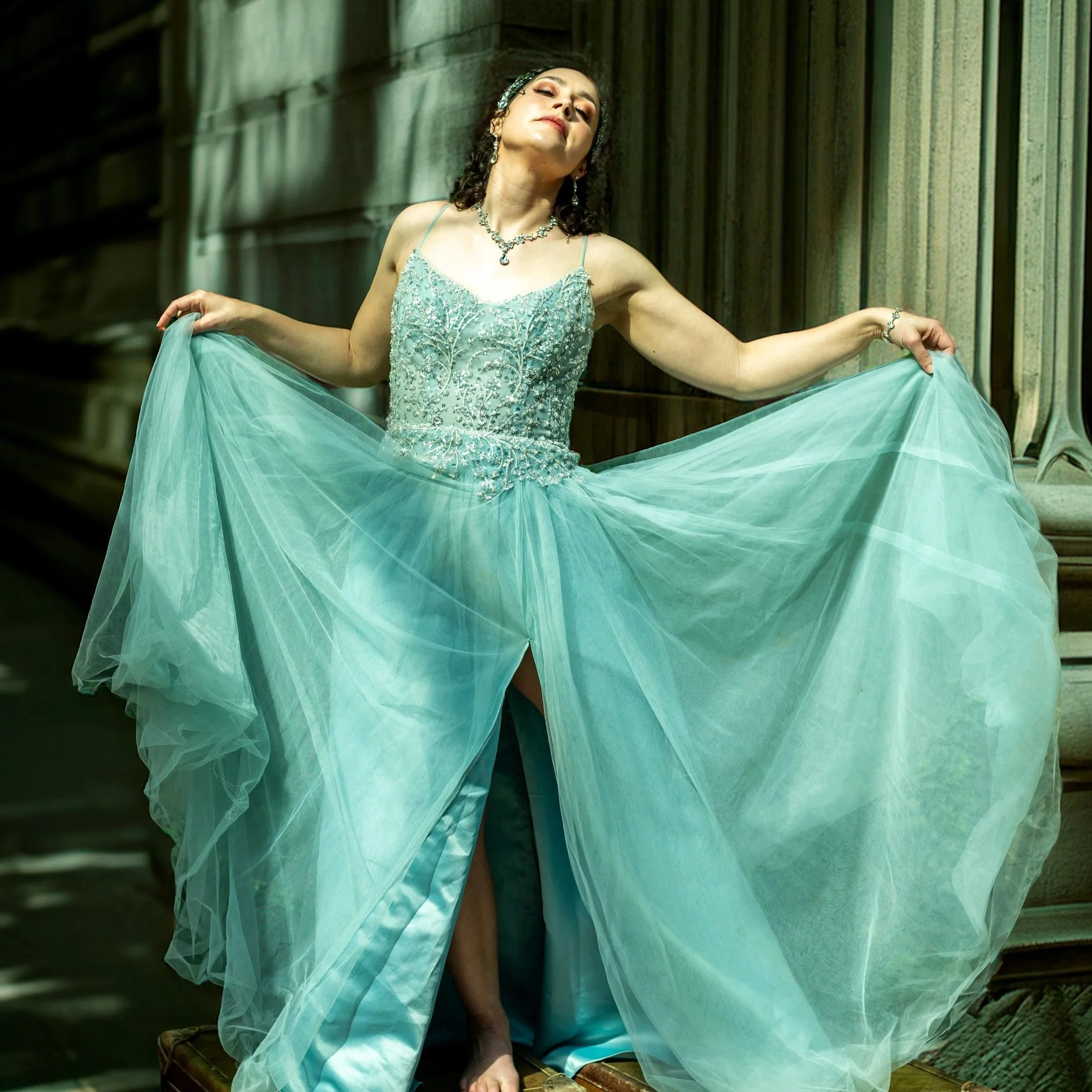 A woman dressed in an elegant, light blue gown with intricate beadwork, standing against a wooden wall with vertical panels. She is holding the fabric of her dress out to the sides, with her eyes closed and head tilted slightly back.