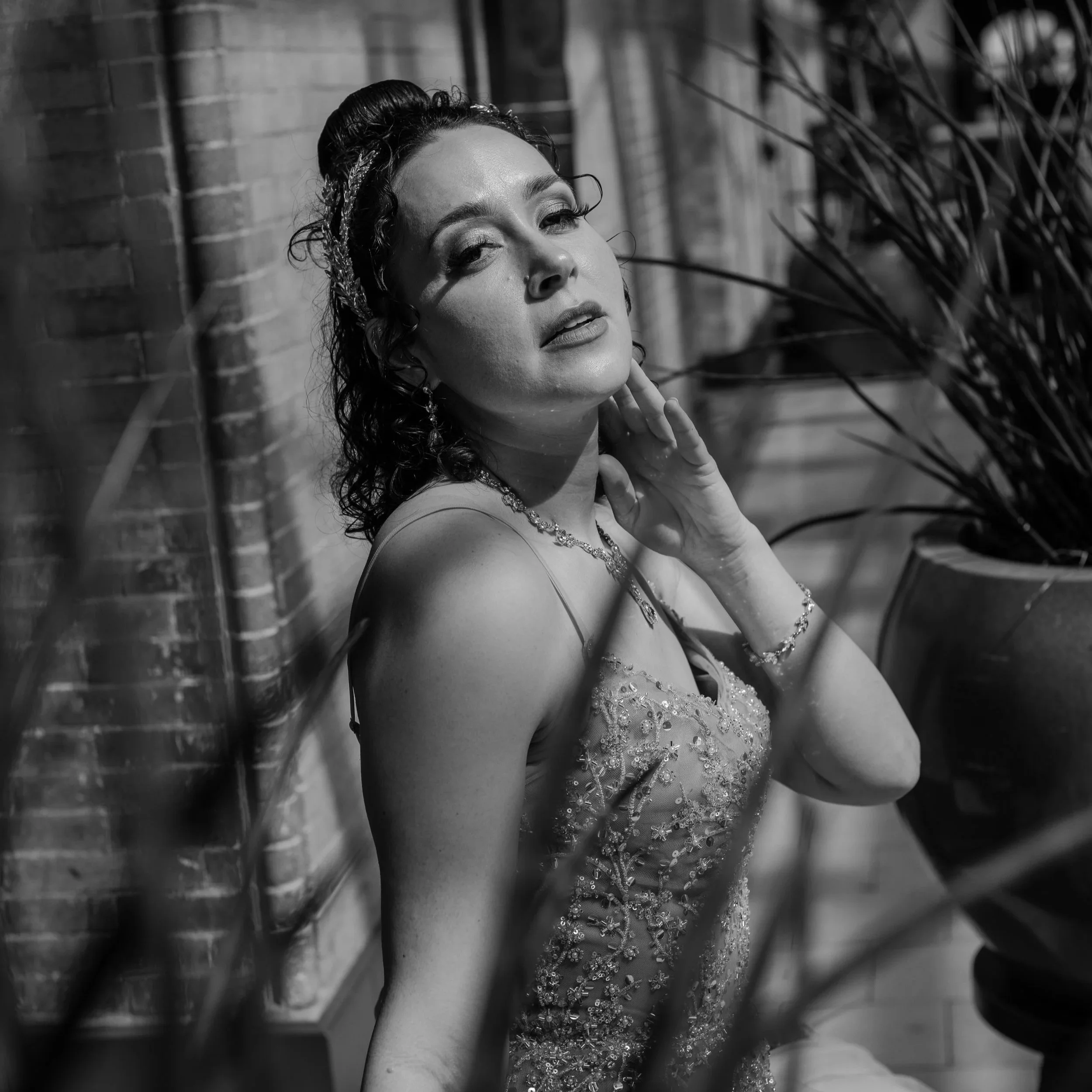 Black-and-white photo of a woman wearing a beaded gown and jewelry, with wet hair styled back, posing against a brick wall with shadows and plants.