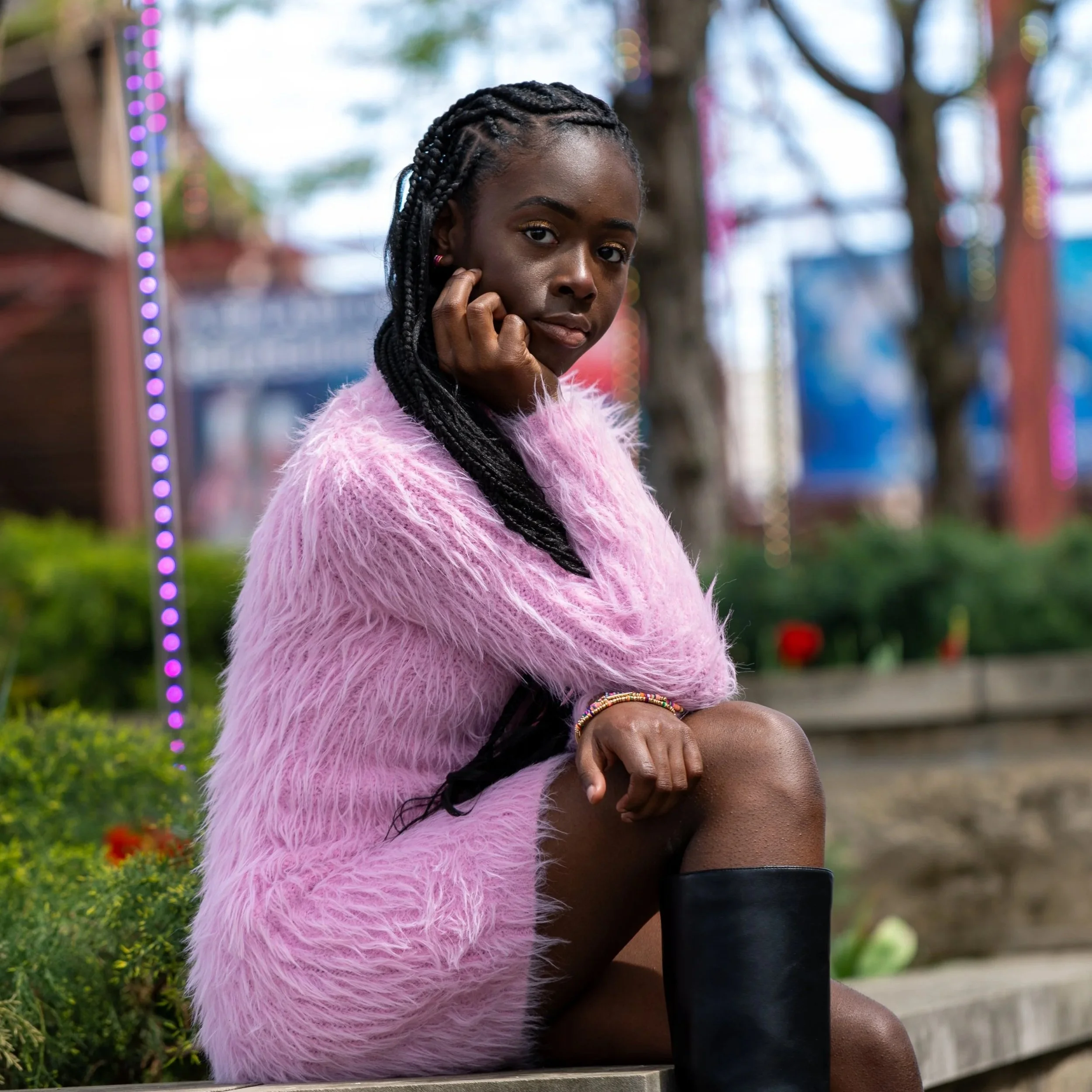 Portrait of a young woman with braided hair, wearing a pink fluffy jacket and sitting outdoors, with colorful lights and trees in the background.