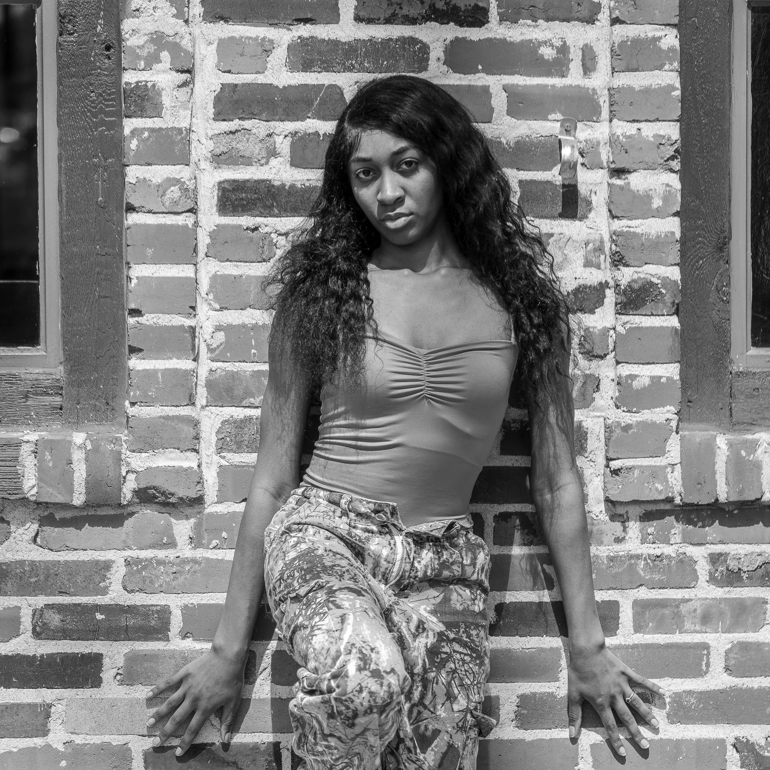 A woman with long curly hair standing against a brick wall, with her hands placed on the wall and her eyes looking at the camera.