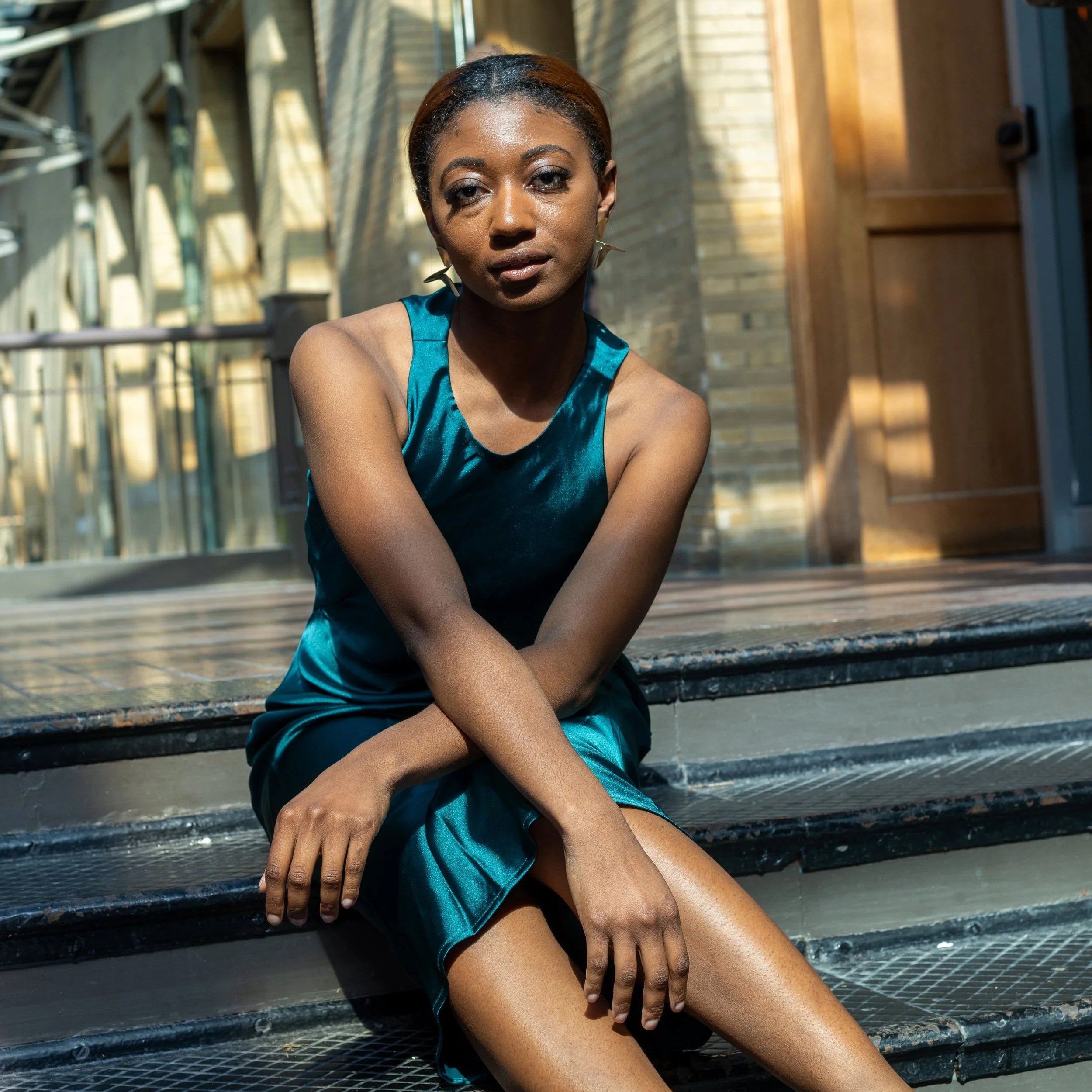 A woman in a teal dress sitting on a metal staircase outdoors, with sunlight and a brick wall background.