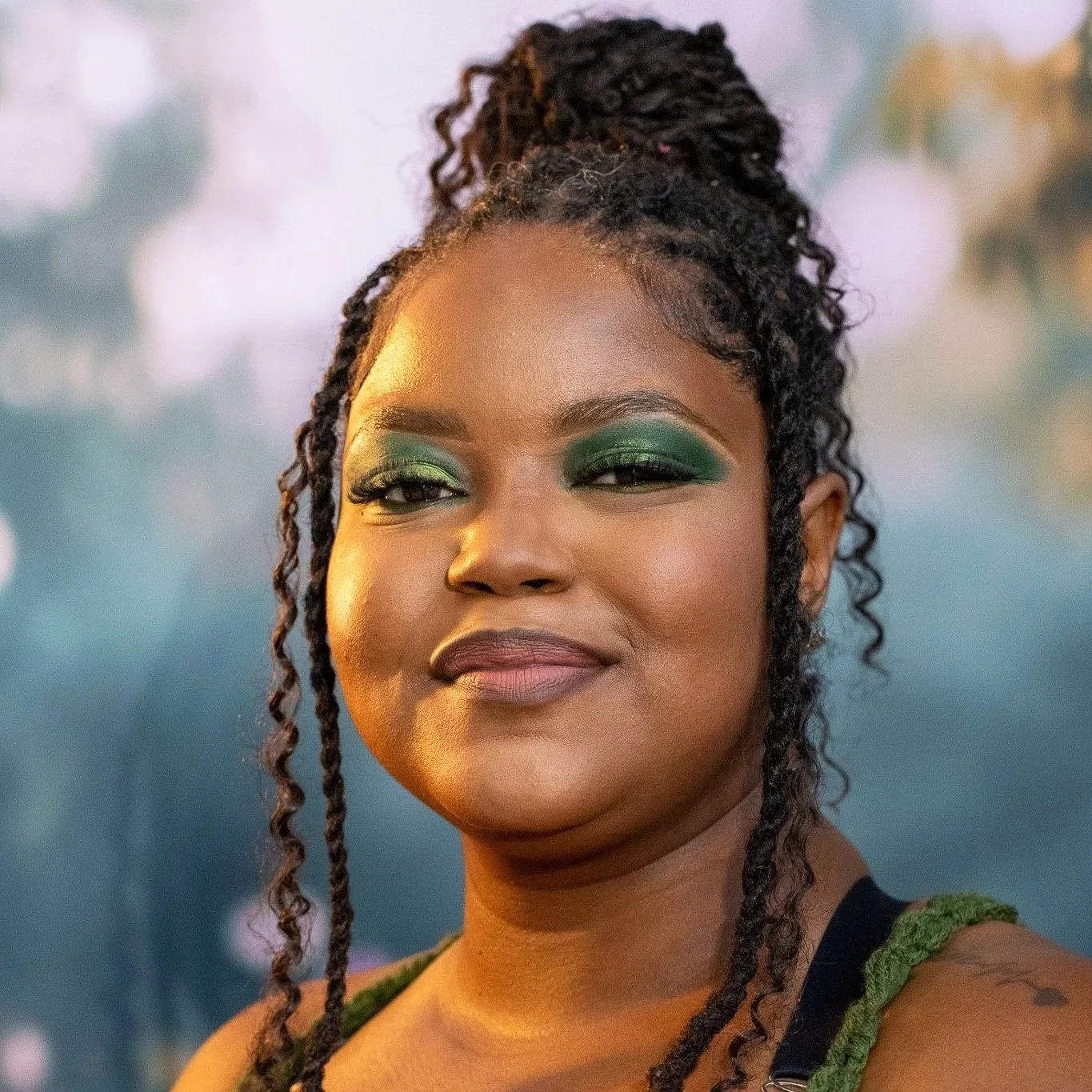 Close-up of a woman with dark skin, wearing green eyeshadow and makeup, with her hair styled in dreadlocks tied up in a high bun, outdoors during the day.