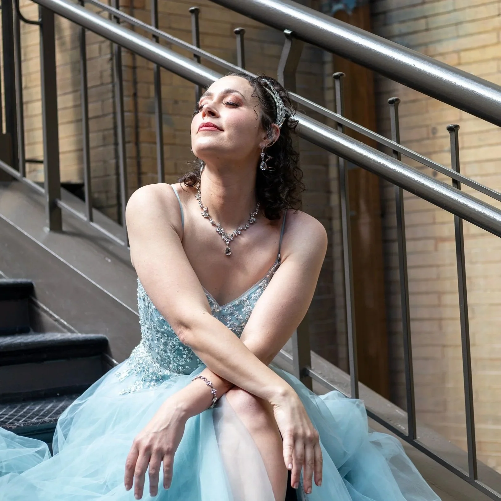 A woman dressed in an elegant, light blue gown with jewelry, sitting on a staircase with her eyes closed and a serene expression.