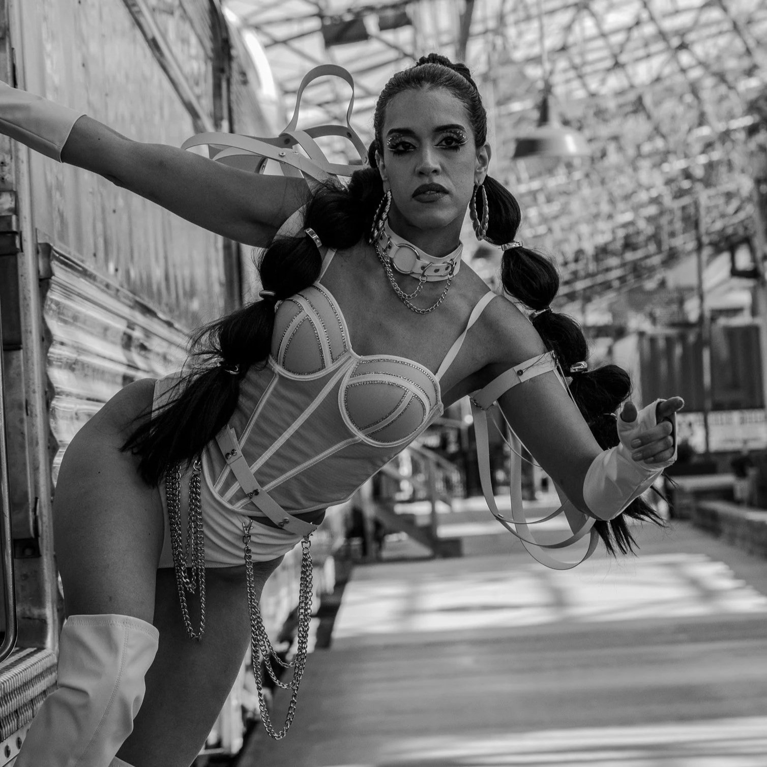 A person in a provocative outfit with pigtails and chains, posing with one arm extended, outside under a glass ceiling structure.