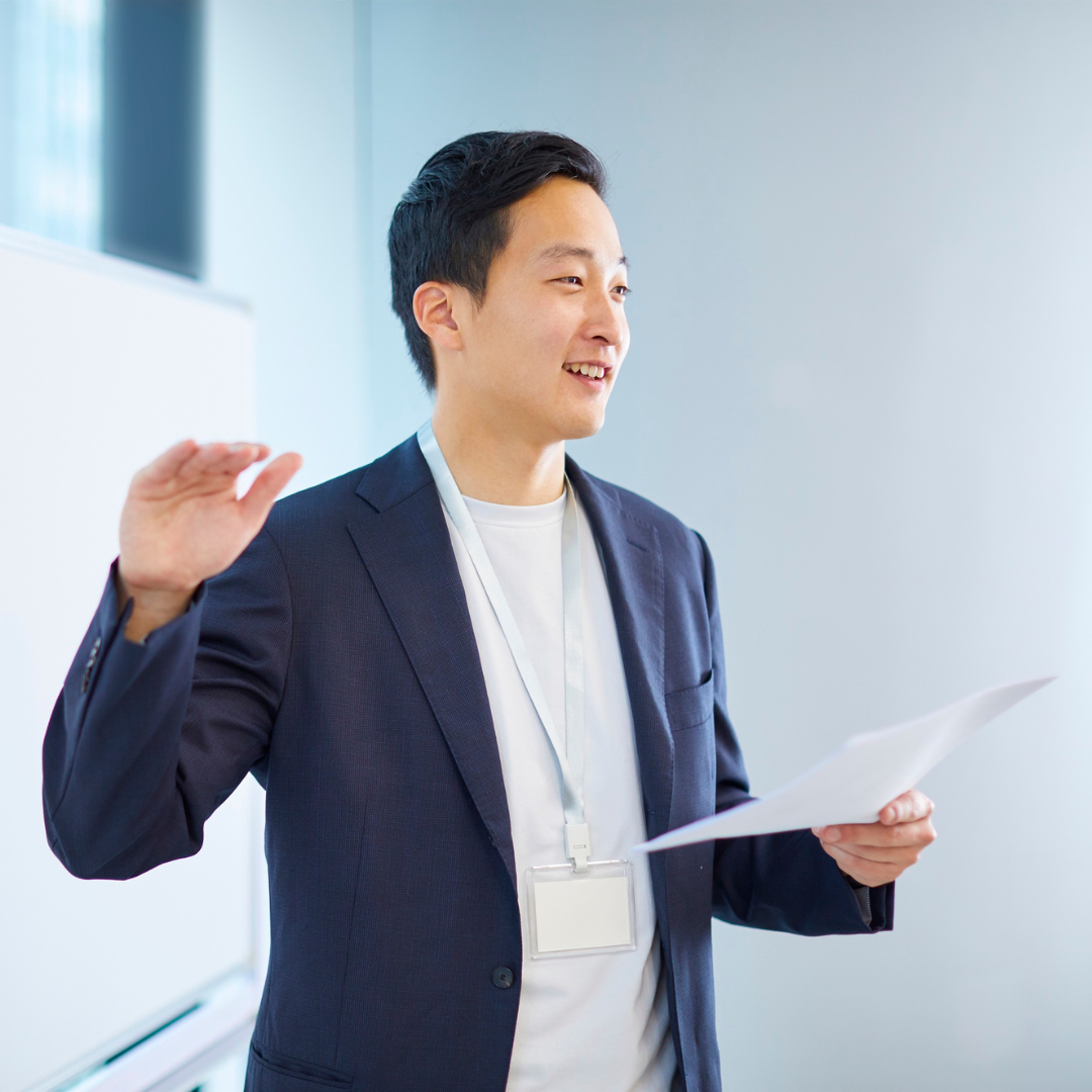 A young man in a navy blazer and white shirt standing in front of a whiteboard, holding papers, smiling, and raising his right hand as if taking an oath or giving a presentation.