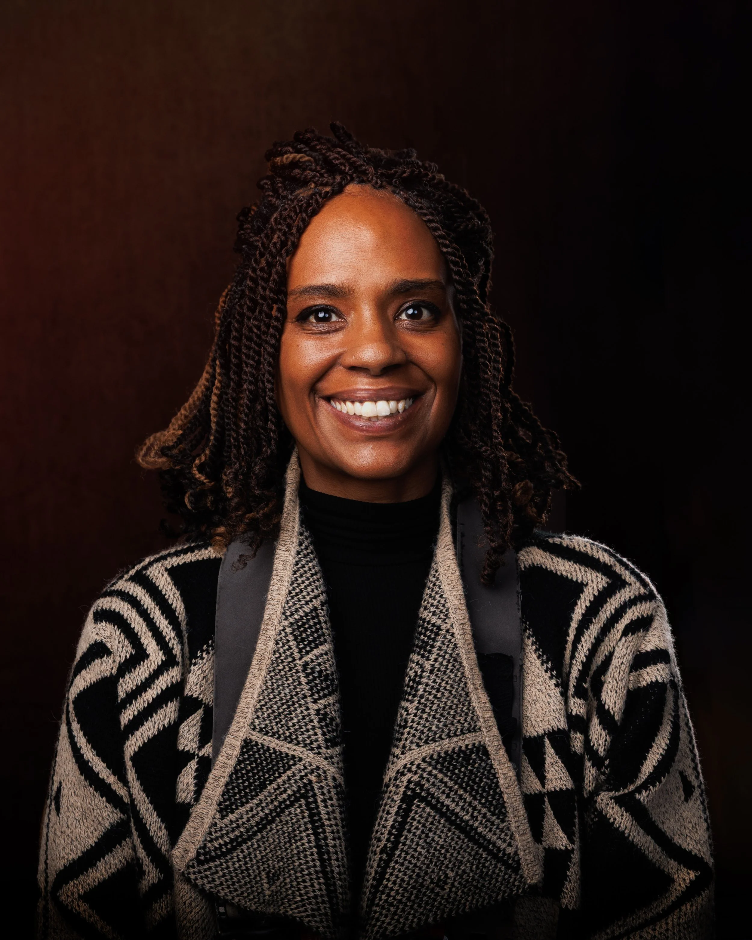 A smiling woman with dark skin and curly hair wearing a patterned coat and a black turtleneck, against a dark background.