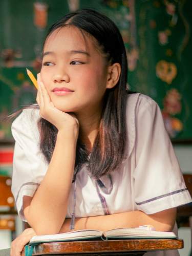 A young girl sitting at a desk with a book open in front of her, resting her chin on her hand, looking to the side with a thoughtful expression, in a classroom setting.