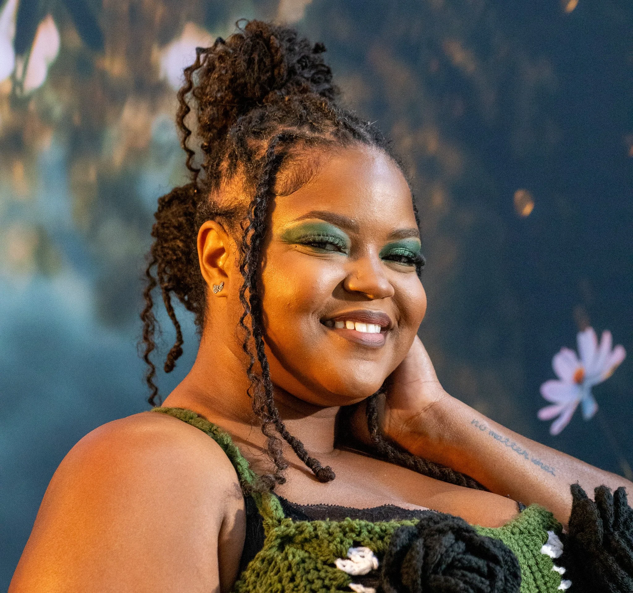 Close-up of a woman with dark brown hair styled in dreadlocks tied up, smiling with her eyes closed, wearing green eyeshadow, earrings, and a green knitted top, with a blue background and blurred floral decorations.