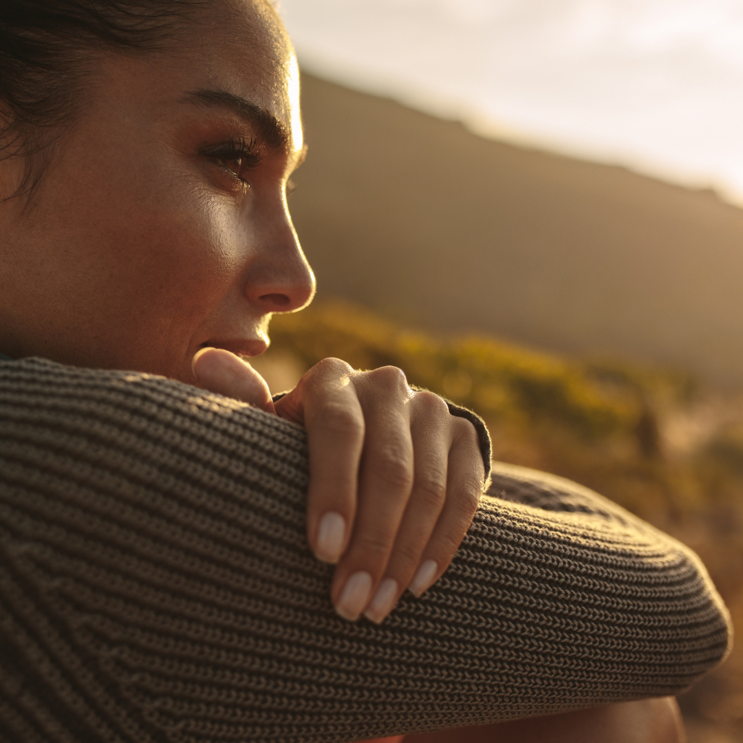 Close-up of a woman lying down outdoors, resting her head on her crossed arms, with a mountain in the background during sunset.