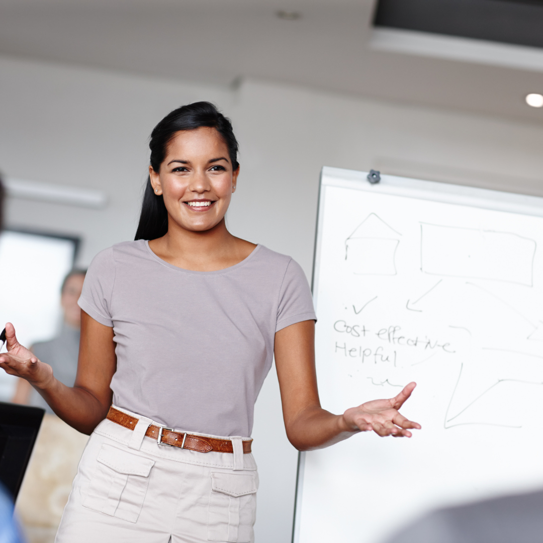 A woman smiling and presenting in front of a whiteboard during a meeting or presentation.