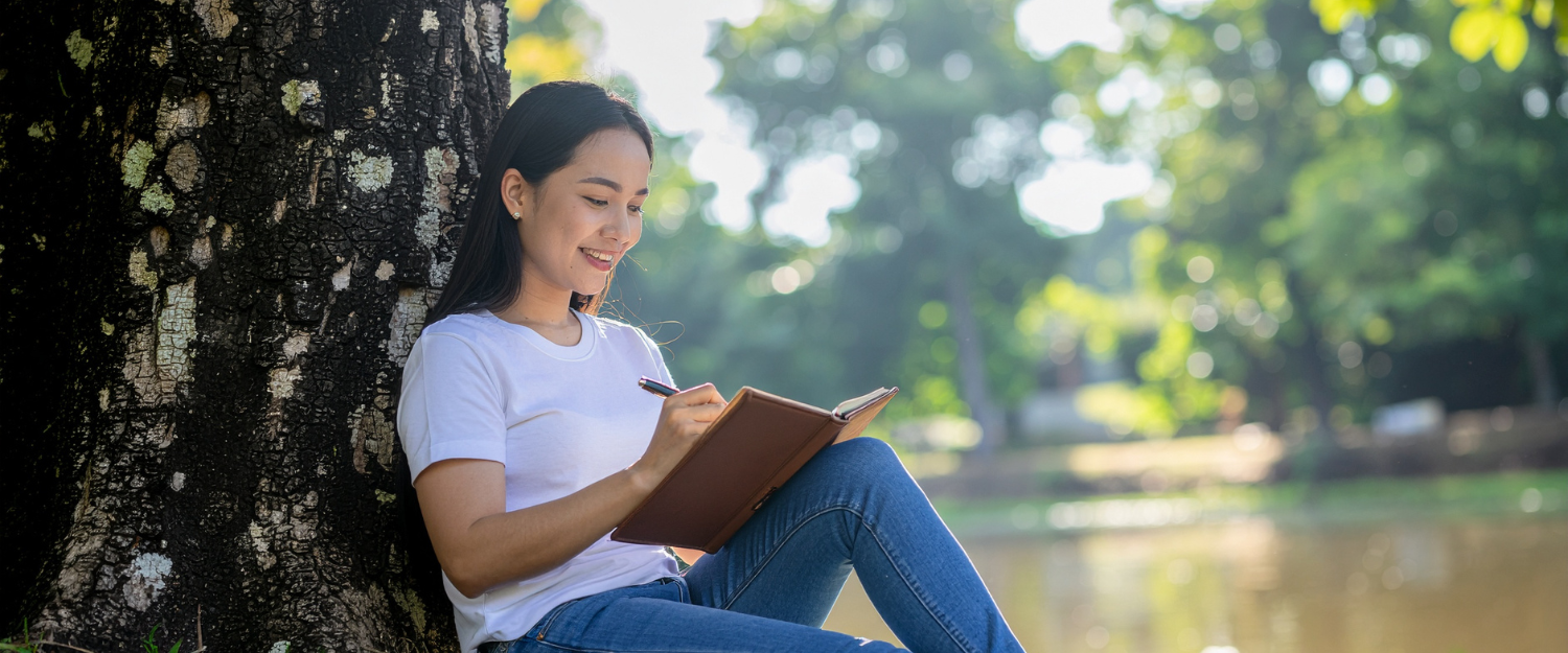Asian woman sitting under a tree journaling in a notebook.