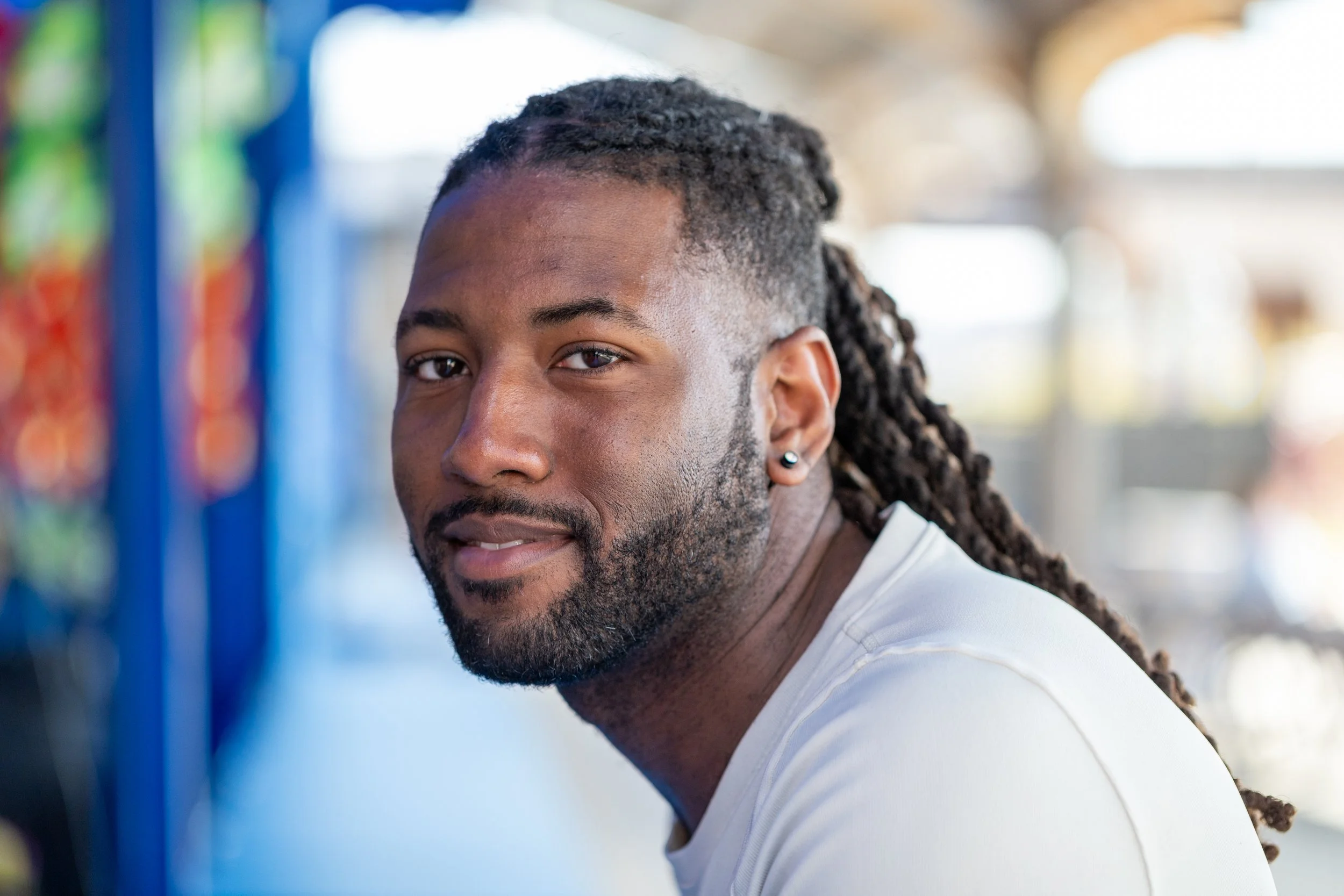 Close-up of a man with dark skin and long dreadlocks, smiling, with a blurred background of colorful objects and outdoor setting.