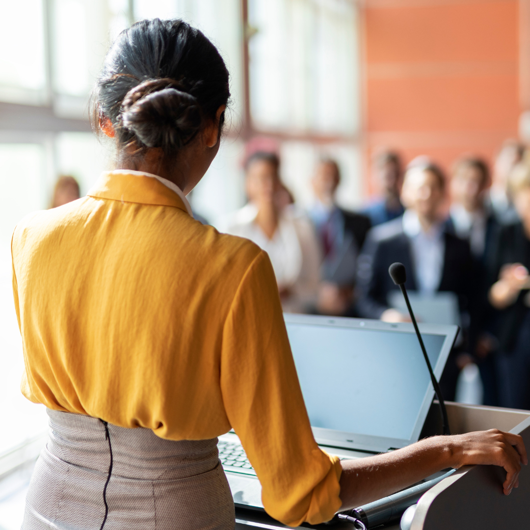 A woman in a yellow blouse giving a presentation to a group of professionals in a conference room.