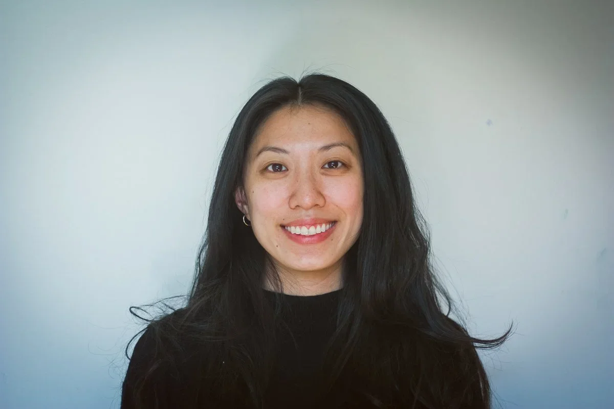 A smiling woman with dark hair, wearing a black top, standing against a plain light-colored wall.