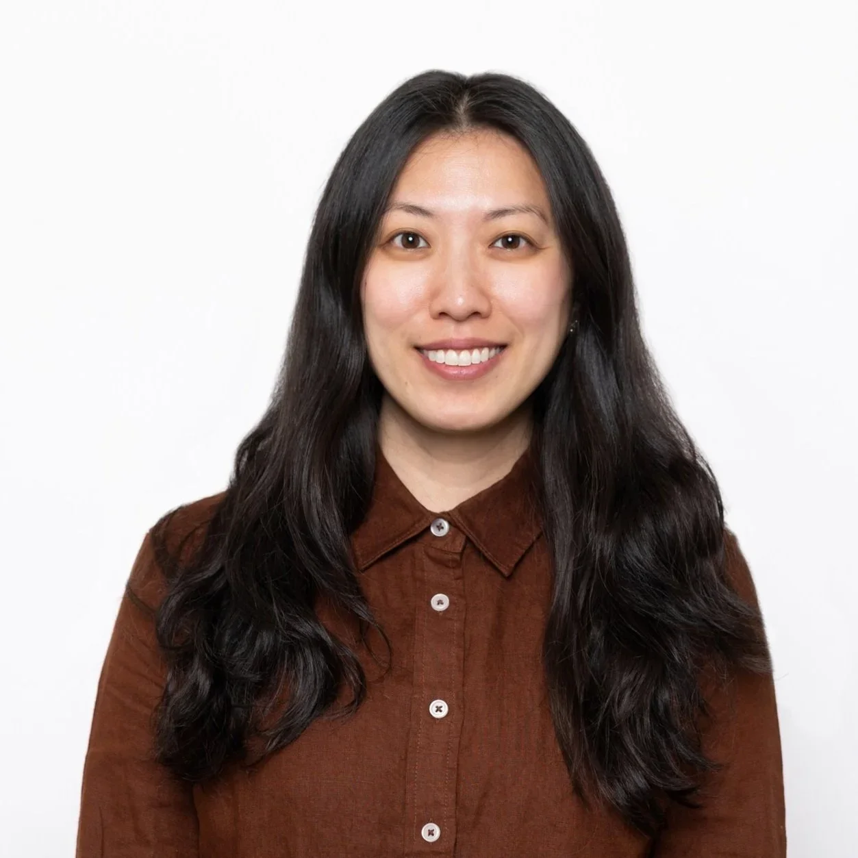 Portrait of a young Asian woman with long dark hair, smiling, wearing a brown button-up shirt against a plain white background.