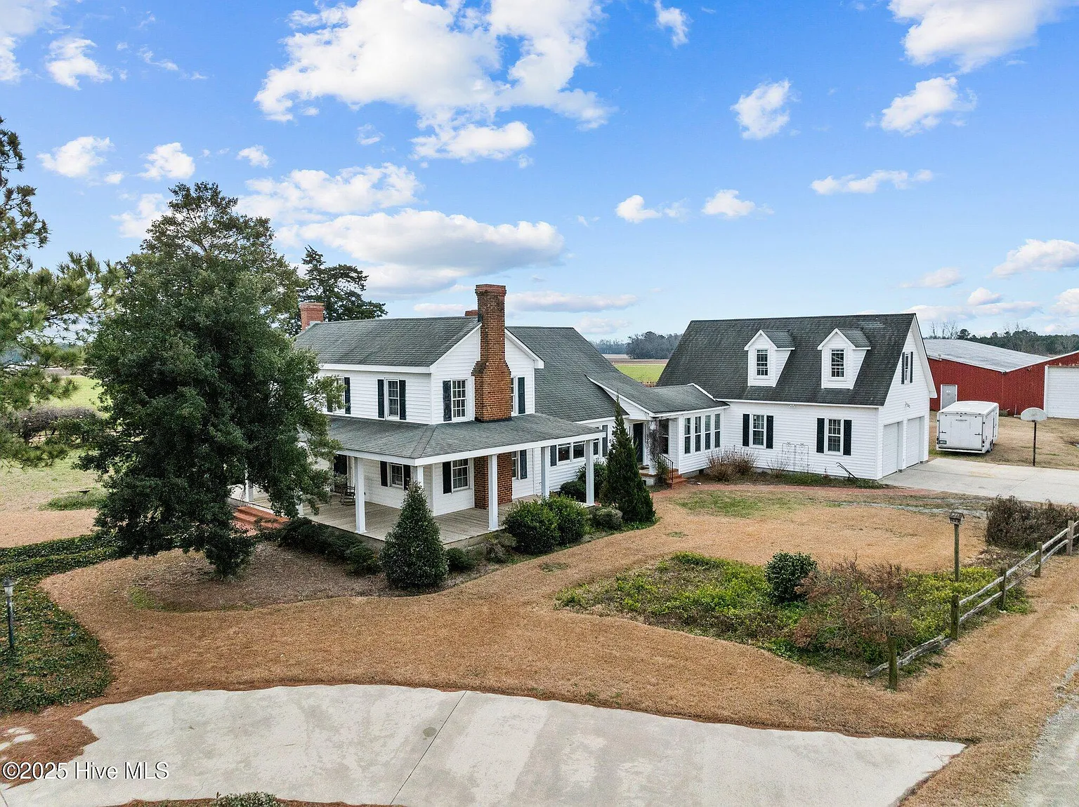 Large white farmhouse-style home with wraparound porch and detached building on rural property under blue sky.