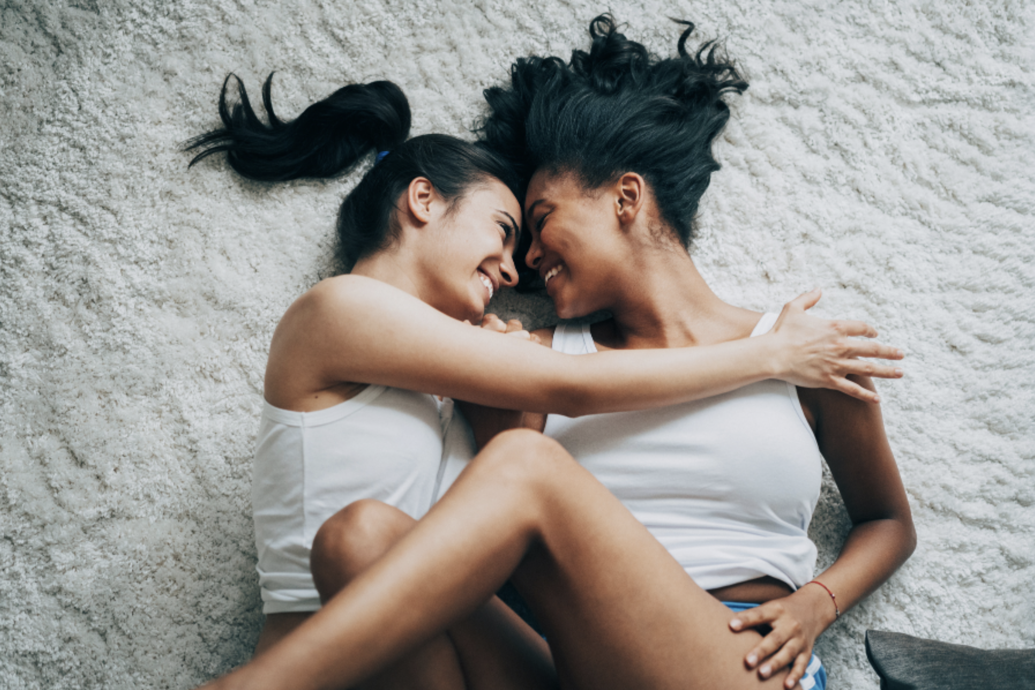 Two women with dark hair lying on a cream shaggy rug, noses touching, smiling, and hugging each other.  Representing emotional intimacy and safety in sex therapy — Attune Therapy Group, Washington State