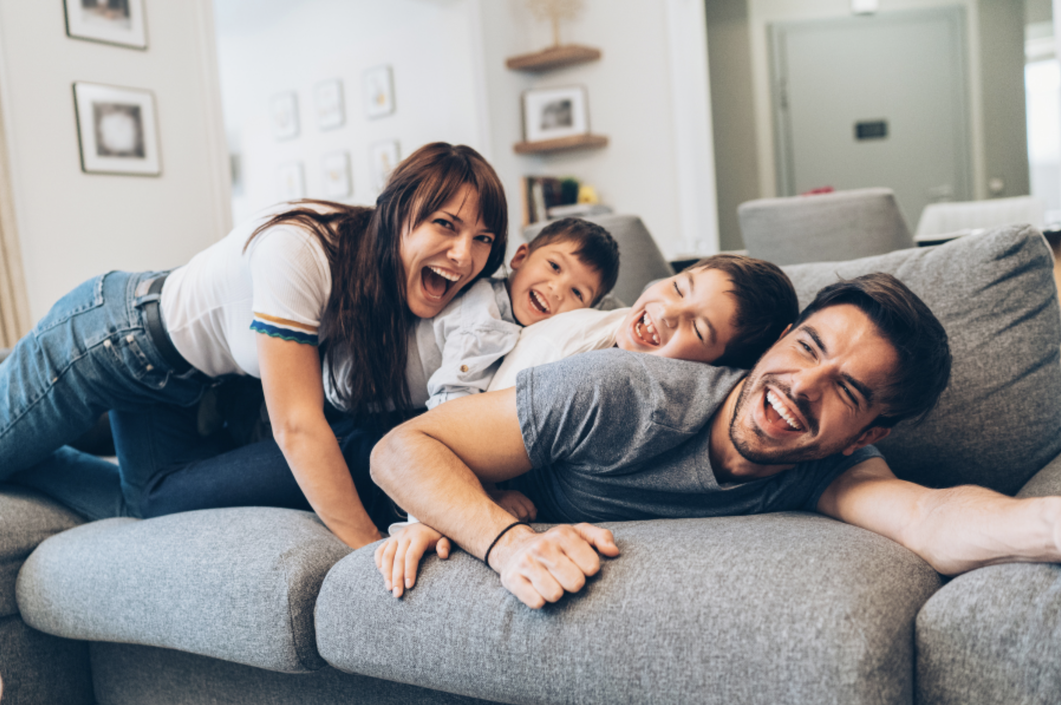 A family of four — mother, father, and two boys — laughing and playing together on a couch, representing the joy and connection that couples therapy for parents can restore — Attune Therapy Group, Washington State
