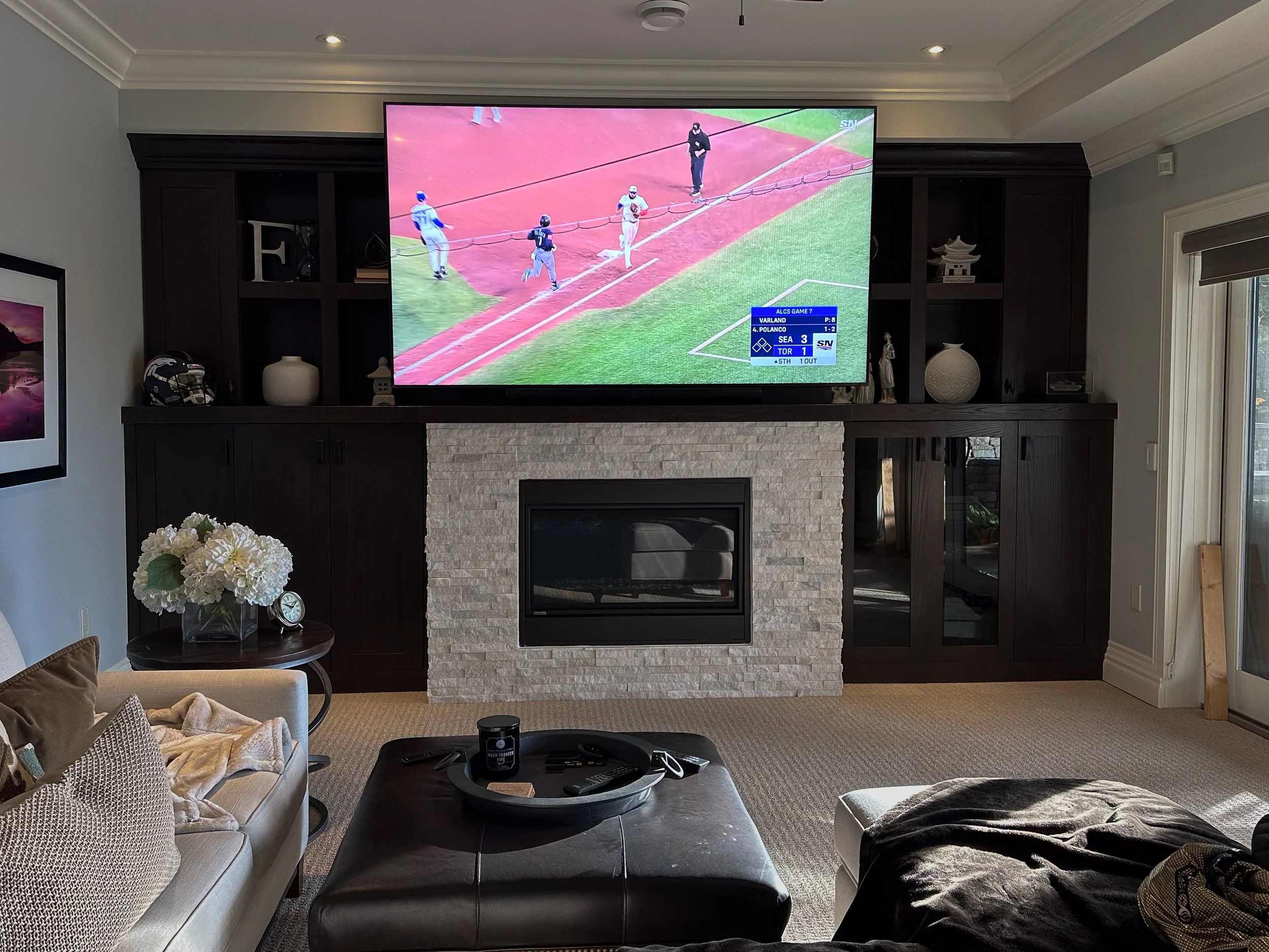 Living room with a large TV showing a baseball game, dark wooden built-in shelves with decorative items, a stone fireplace, a beige sofa with pillows, a black ottoman with a tray and remote controls, a side table with white flowers and a clock, and a