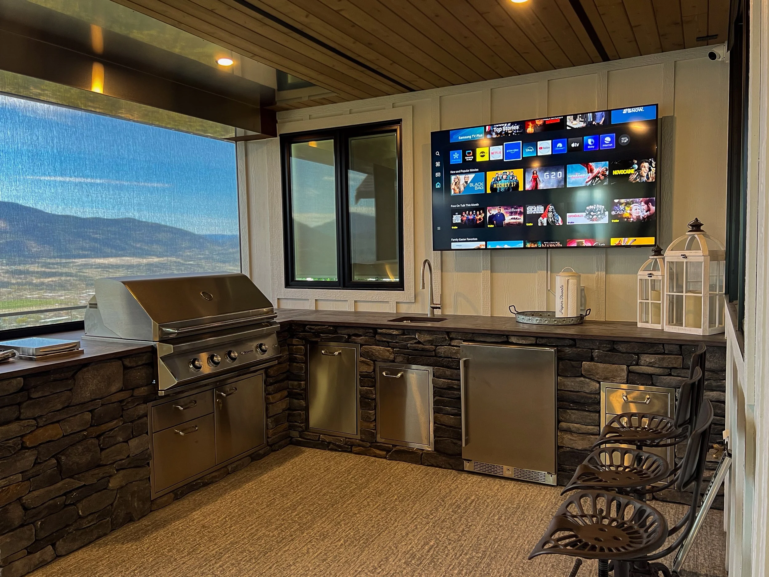 Outdoor patio with a stone countertop, stainless steel grill, three black chairs, a large TV mounted on a white wall, with mountainous scenery visible through large windows.