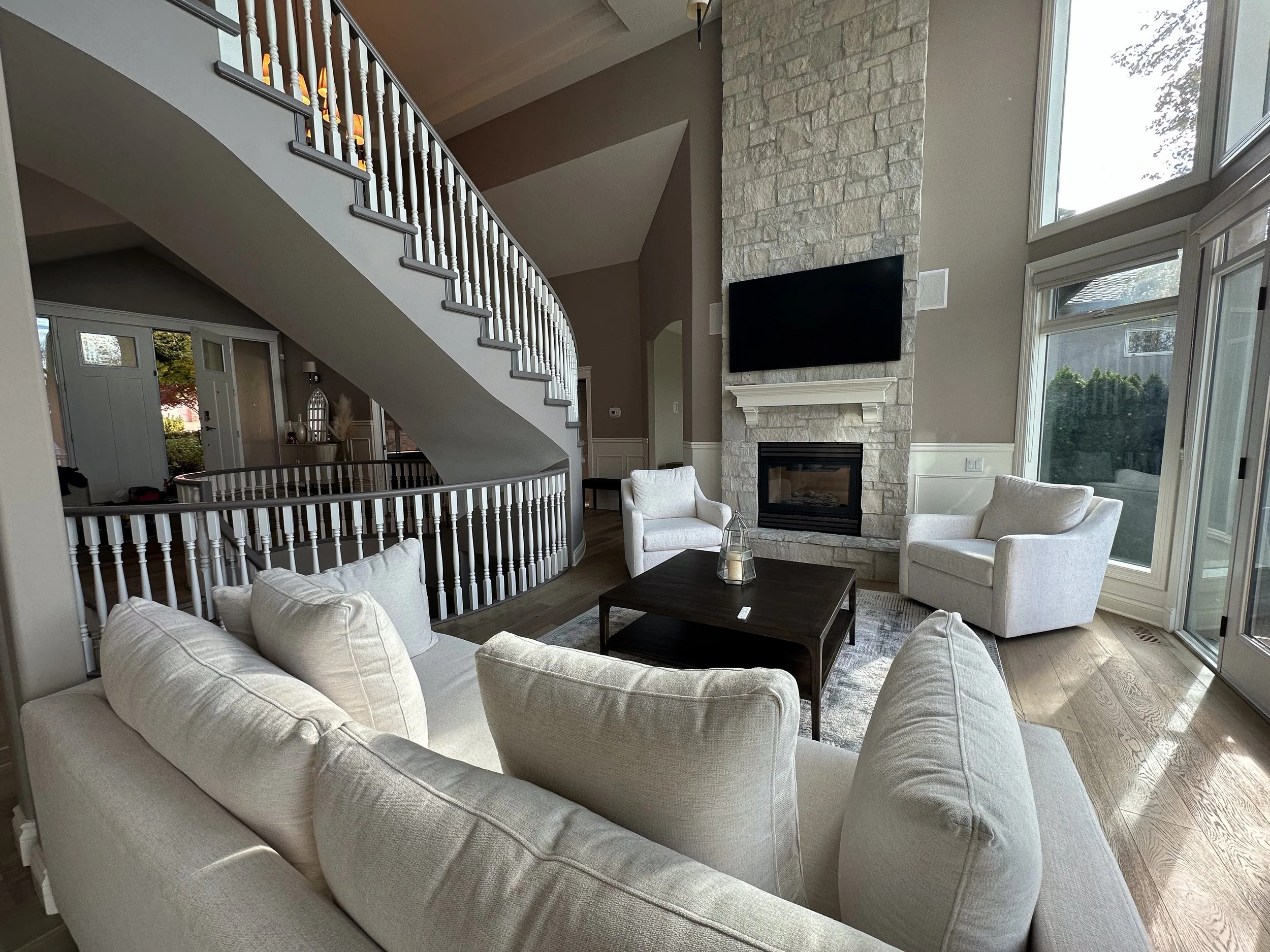 Living room with white sofa, two white armchairs, a dark wood coffee table, a fireplace with a stone surround, and a black mounted television above the fireplace. Large windows on the right side let in natural light.