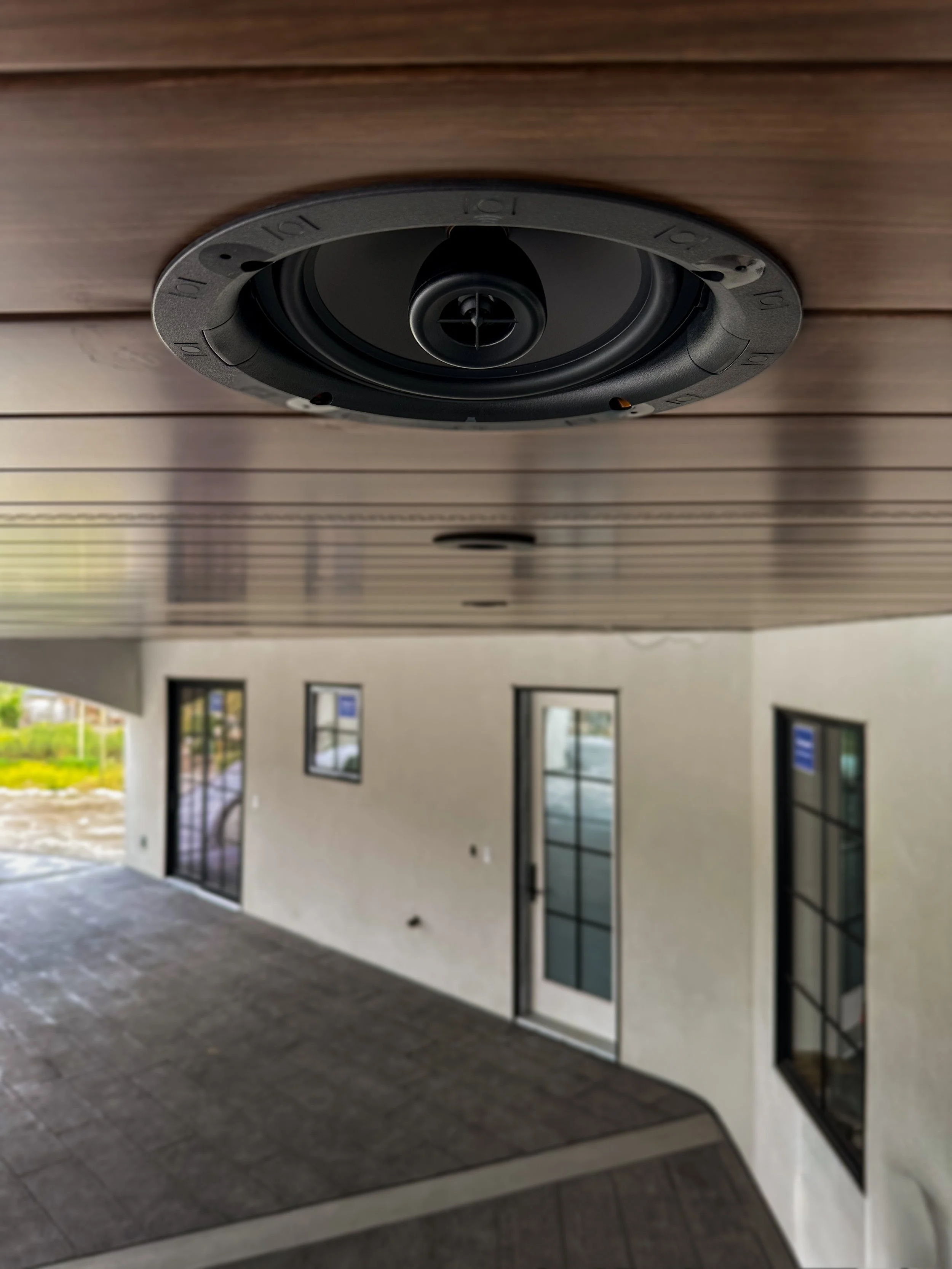 Close-up of a black round ceiling speaker installed under a wooden ceiling in an outdoor patio area, with a house wall and windows in the background.