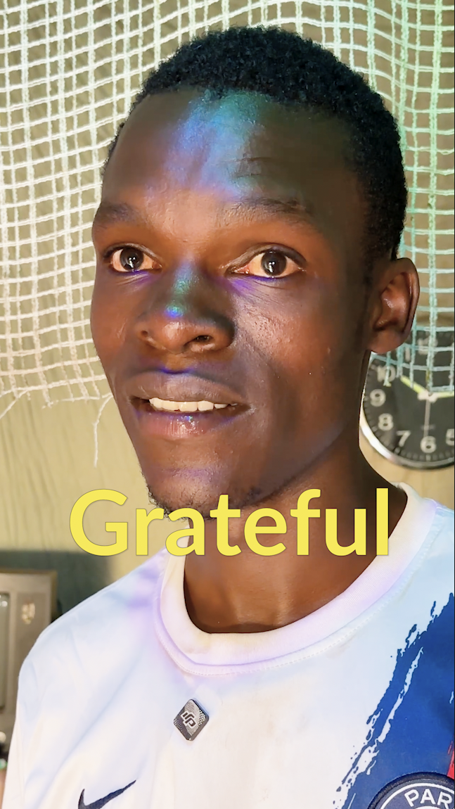 A young man with short black hair and a slight smile, standing in front of a sports net and a wall clock, wearing a white sports jersey with blue and red details, with the word 'Grateful' written in large yellow letters across the front.