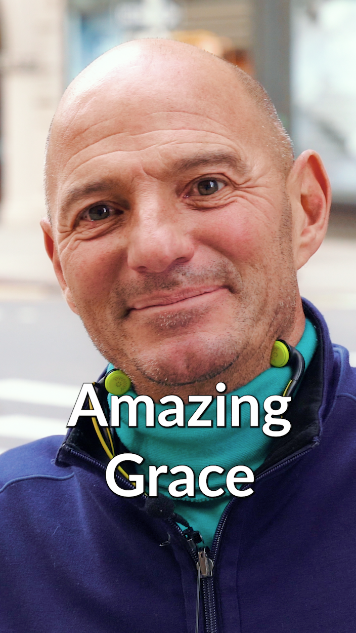 Close-up of a smiling bald man wearing a blue jacket and green wireless earbuds, with the text 'Amazing Grace' overlaid.