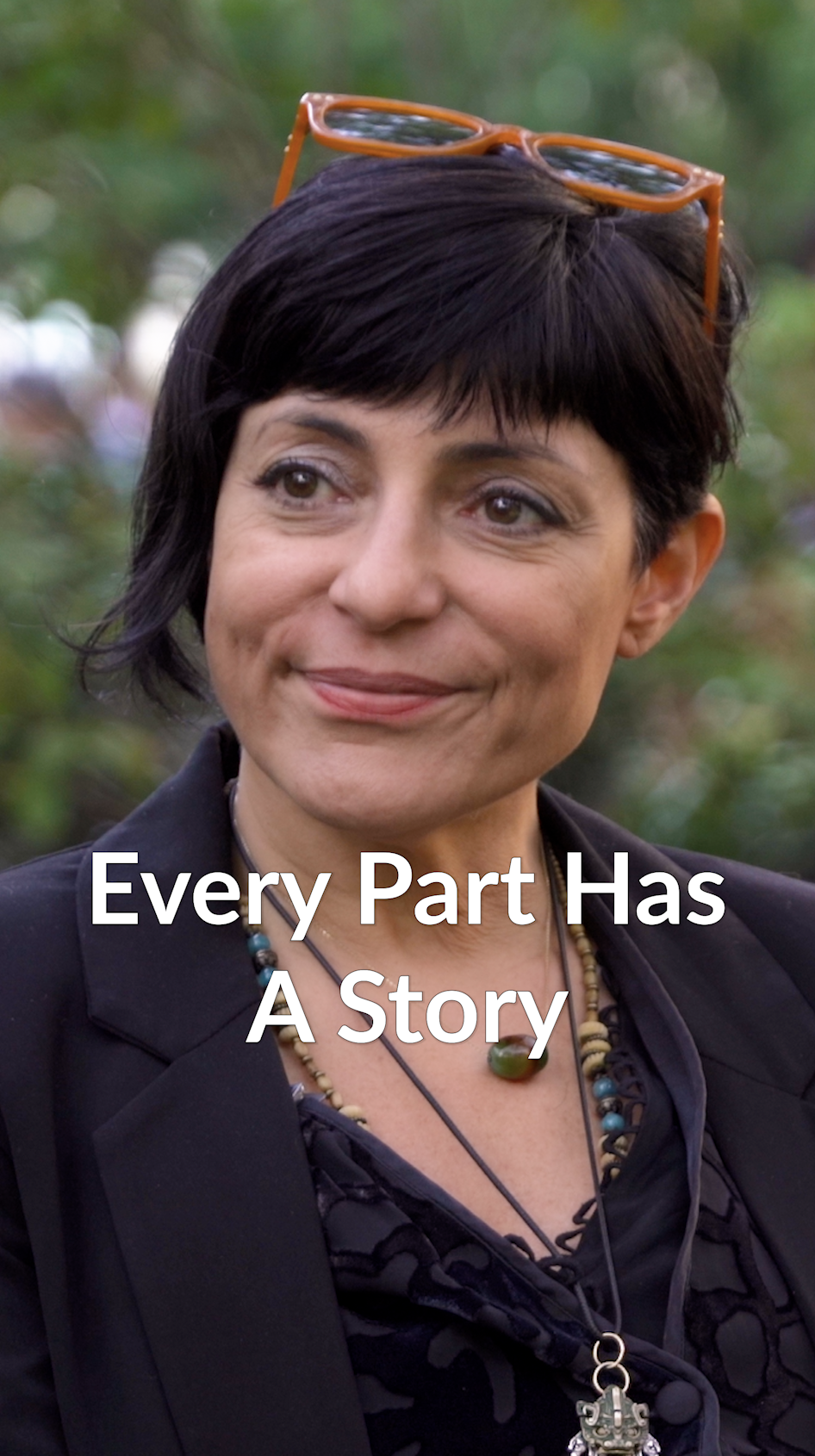 A woman with short black hair wearing sunglasses on her head, a black blazer, and layered necklaces, outdoors with a blurred green background, smiling with the text 'Every Part Has A Story' superimposed.