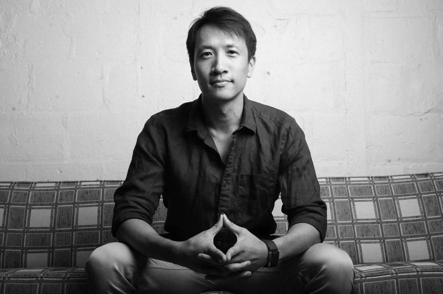 A young man with short dark hair, sitting on a patterned couch against a plain wall, with his hands clasped together in a relaxed pose, wearing a long-sleeve shirt and a wristwatch.