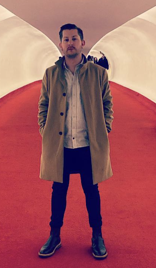 A man stands in an architectural setting with white and red curved walls, wearing a beige coat, white shirt, dark pants, and black boots.