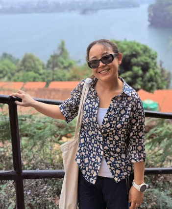 Woman wearing sunglasses and a patterned shirt, smiling, standing outdoors by a railing with a lake and trees in the background.